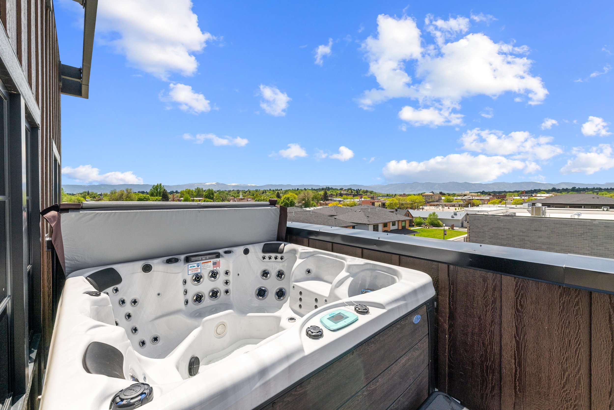 Hot tub on a balcony overlooking a suburban neighborhood under a bright blue sky with clouds.
