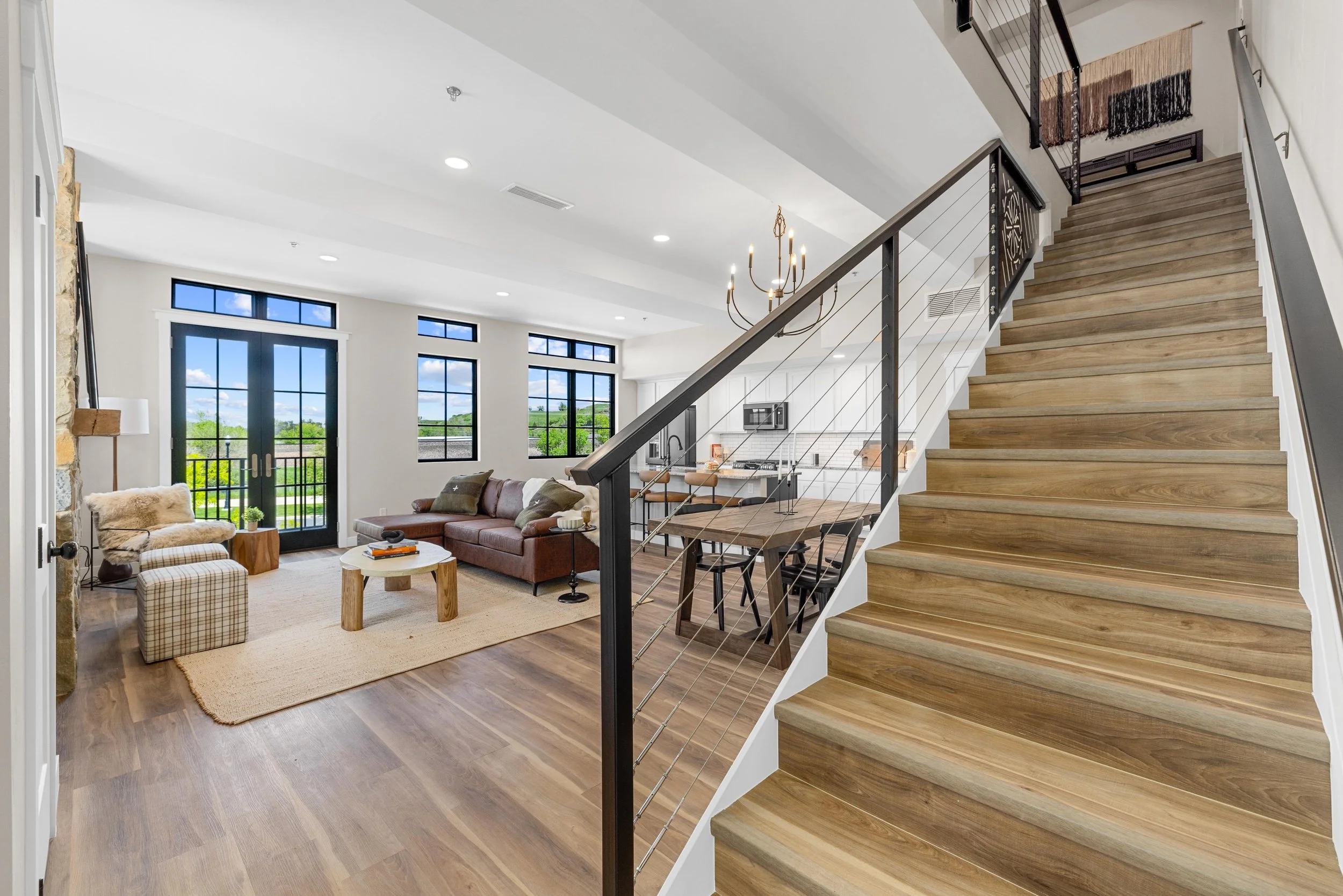 Open living room with large windows, brown sofa, fireplace, wooden coffee table, and stairs leading upstairs.