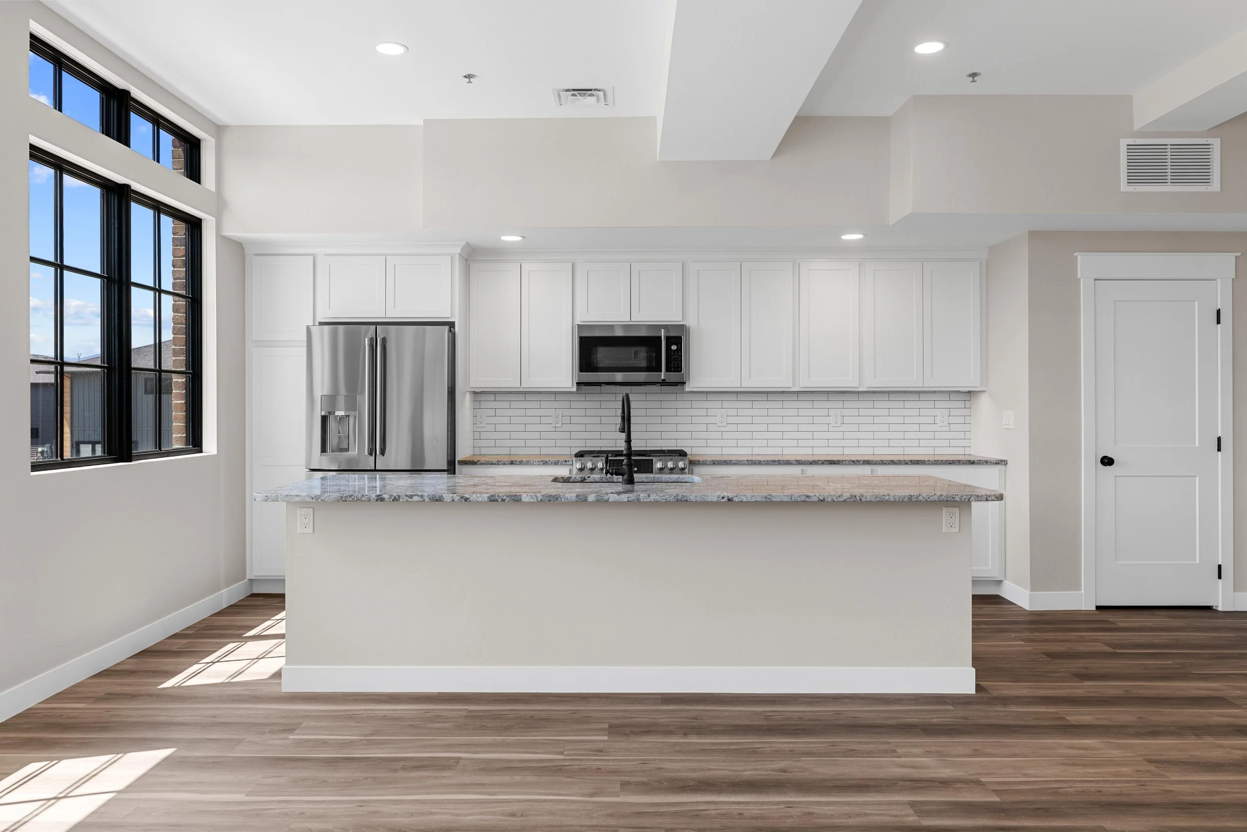 Modern kitchen with white cabinets, a granite countertop island, stainless steel refrigerator, microwave, black faucet, and white subway tile backsplash, illuminated by recessed lighting with a large window.