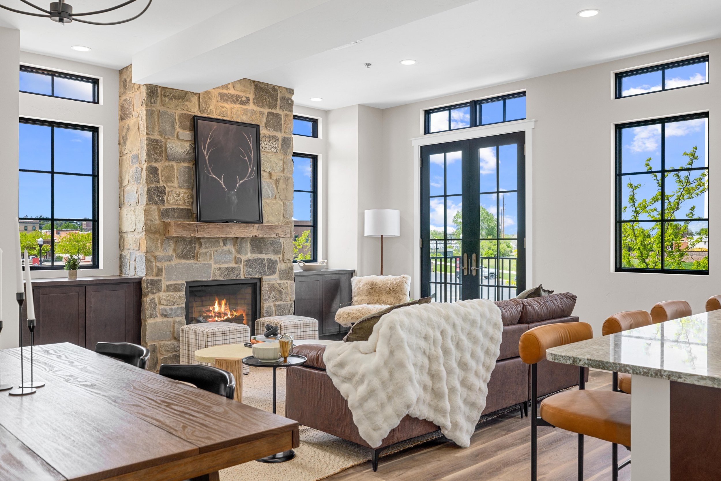 Modern living room with a stone fireplace, large windows, a brown sofa with a white fluffy blanket, and a sliding glass door leading outside.