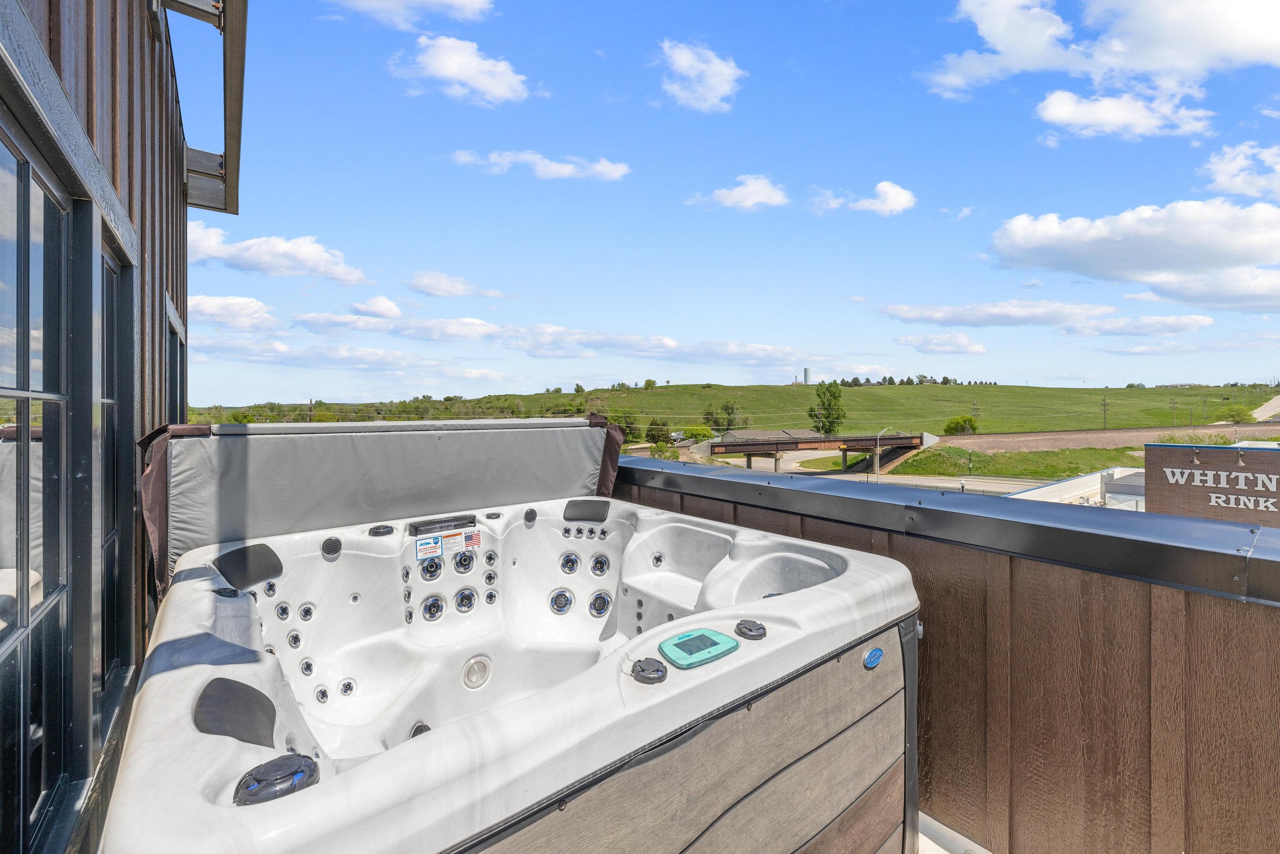 Outdoor hot tub on a balcony overlooking a grassy landscape with blue sky and scattered clouds.
