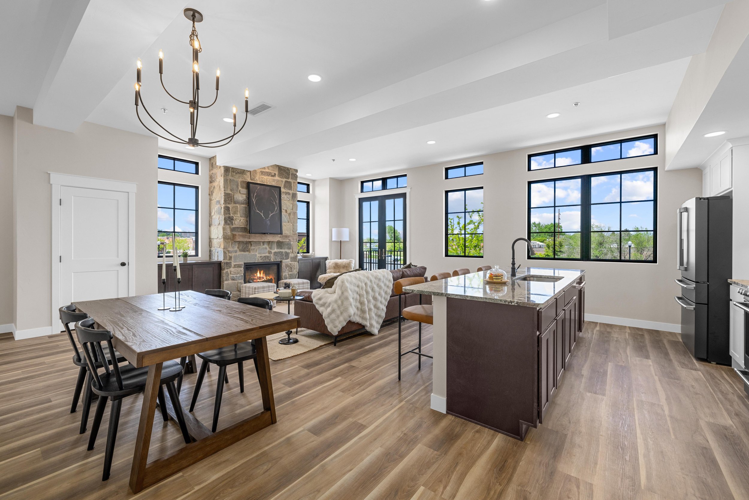 Open-concept living room and kitchen with large windows, stone fireplace, fireplace, dining table, bar stools, modern chandelier, and hardwood floors.
