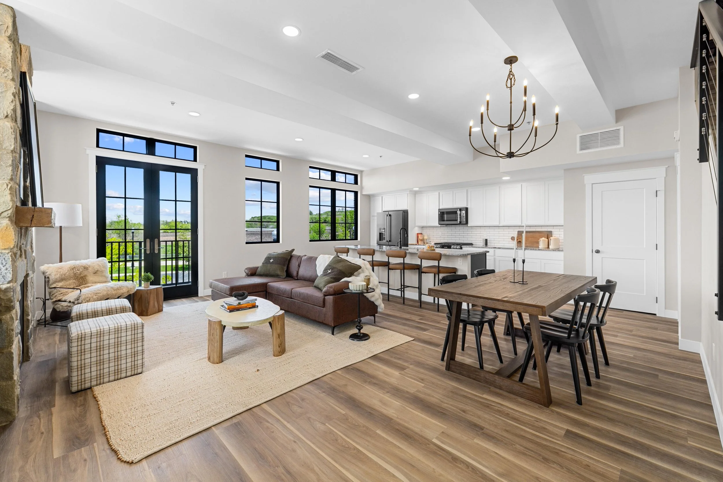 Open-concept living room and kitchen with large windows, a brown sofa, a white kitchen, a wooden dining table, and a chandelier.