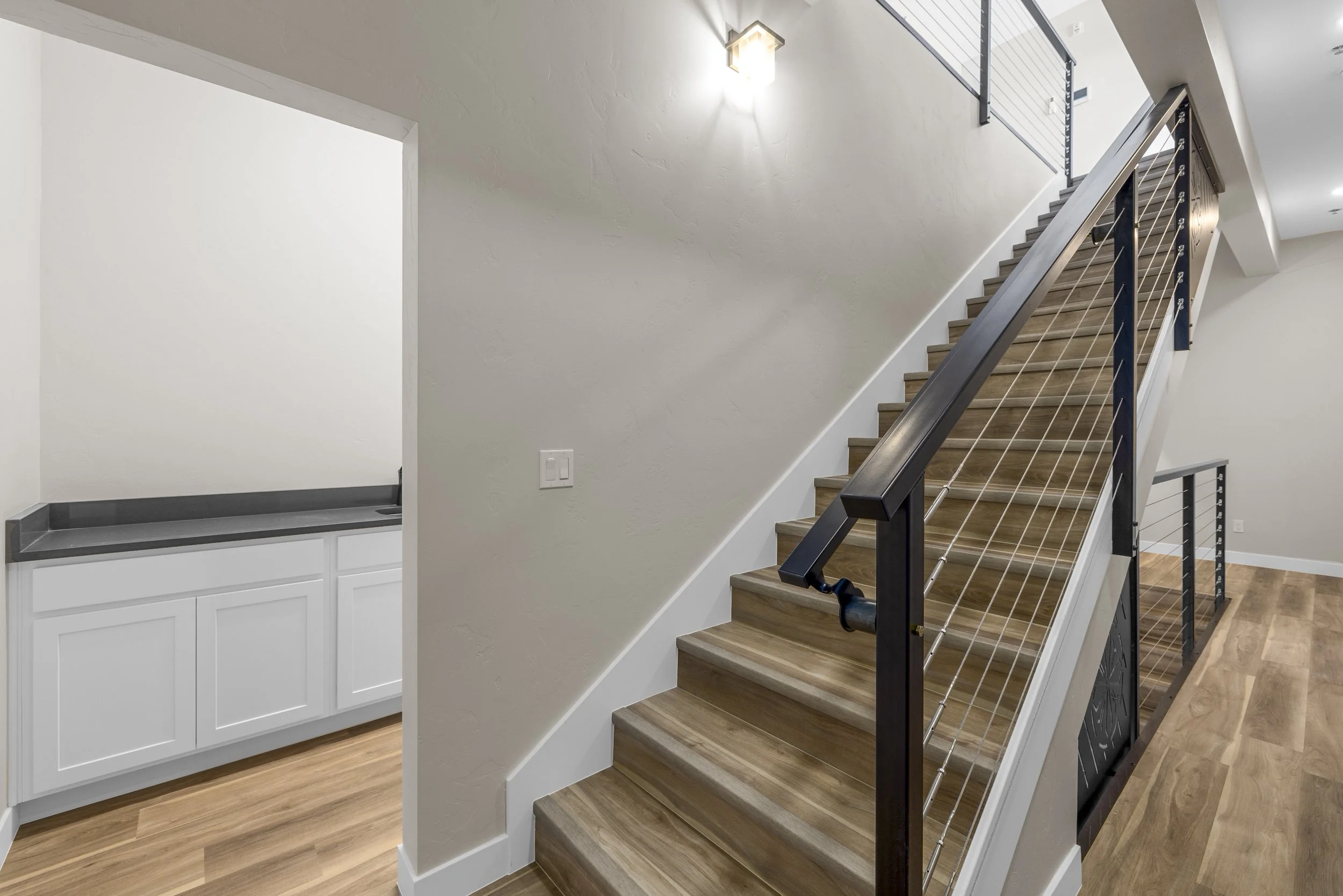 Interior view of a modern home showing a staircase with wooden steps and a black metal handrail, a wall-mounted light fixture, and a partial view of a laundry area with white cabinets and a gray countertop.