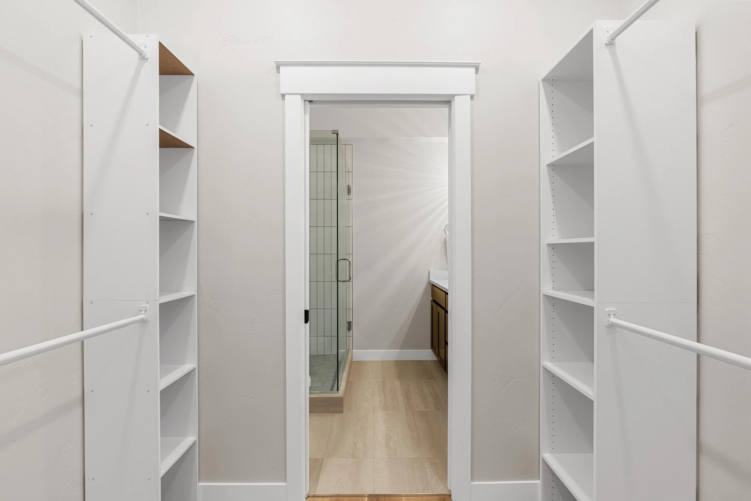 Empty walk-in closet with white shelves and rods, leading to bathroom with tiled shower and wooden vanity.