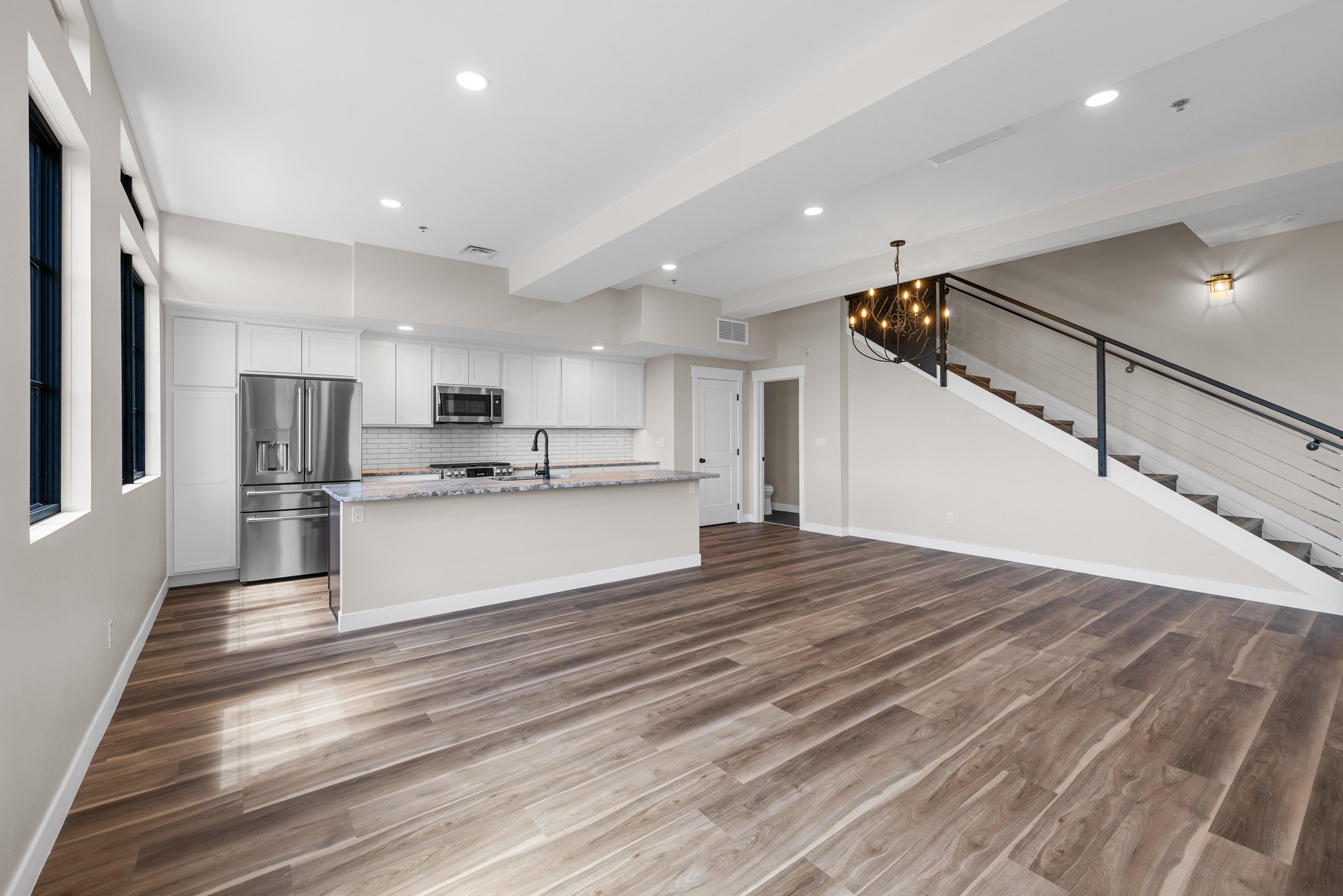 Open-concept living space with kitchen, hardwood floors, staircase, and modern lighting fixtures.