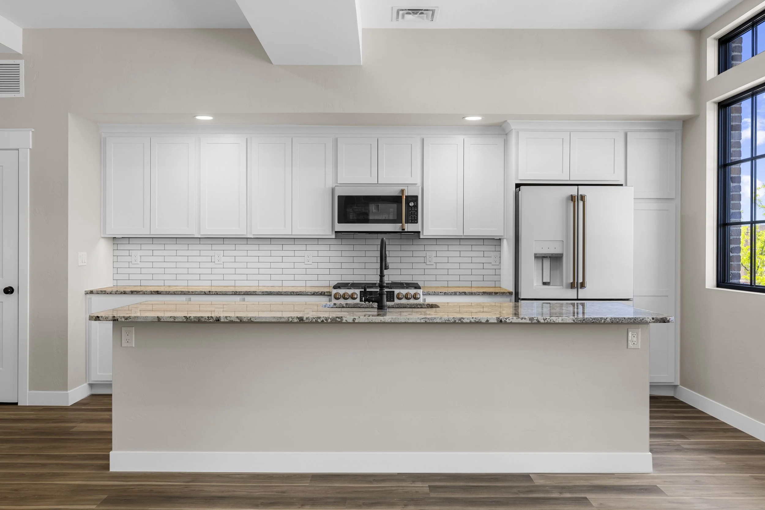 Modern kitchen with white cabinets, a granite countertop island, a stainless steel refrigerator with double doors, an oven, a microwave, and a backsplash with white subway tiles. Large windows on the right provide natural light.