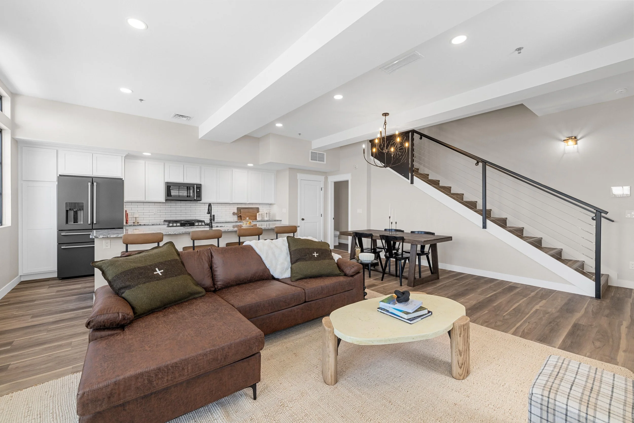 Open-concept living room and kitchen with white cabinets, stainless steel refrigerator, brown sofa, wooden coffee table, and staircase with spa curtain, modern lighting, and wood flooring.