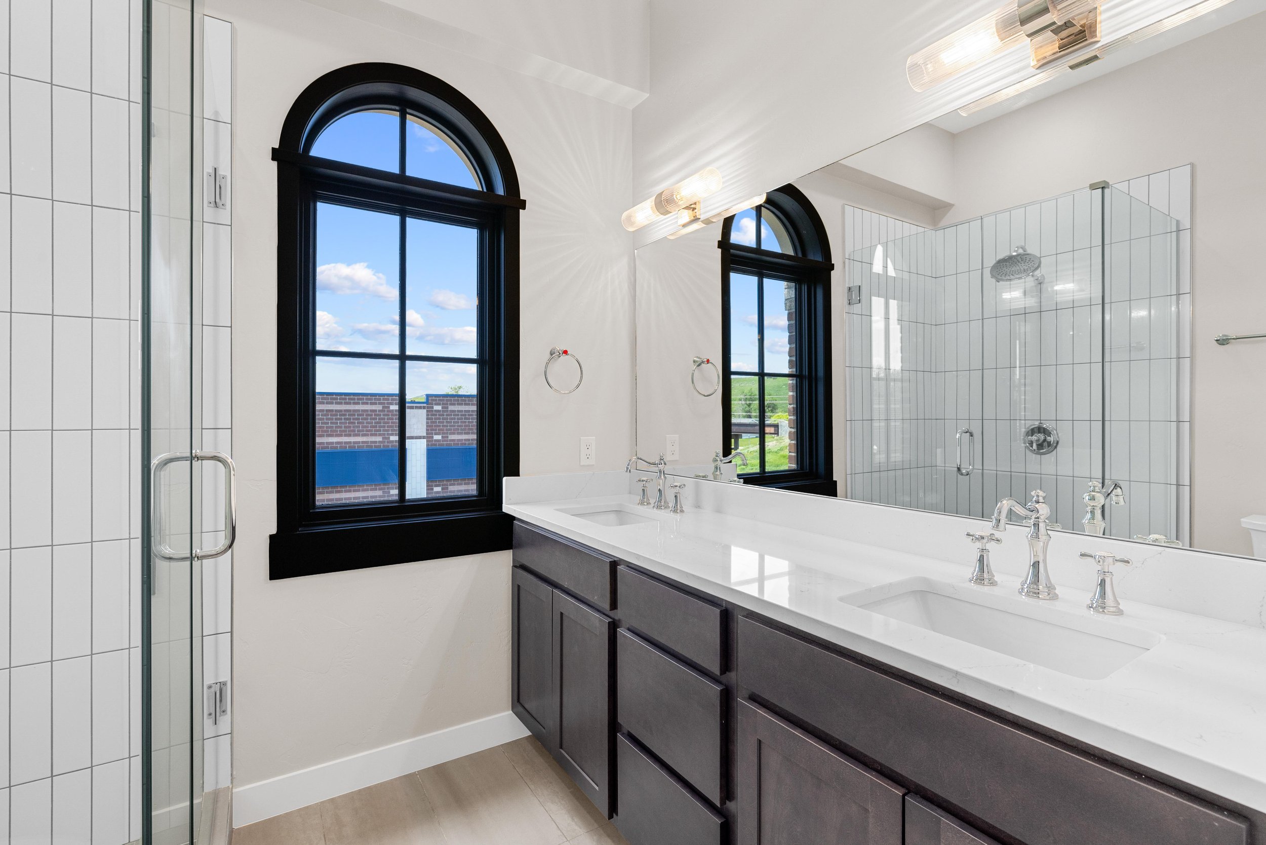 Modern bathroom featuring a large white double vanity with gray cabinets, a big mirror, black framed arched windows showing a blue sky with clouds, a glass-enclosed shower with a rain showerhead, and light wood floor tiles.