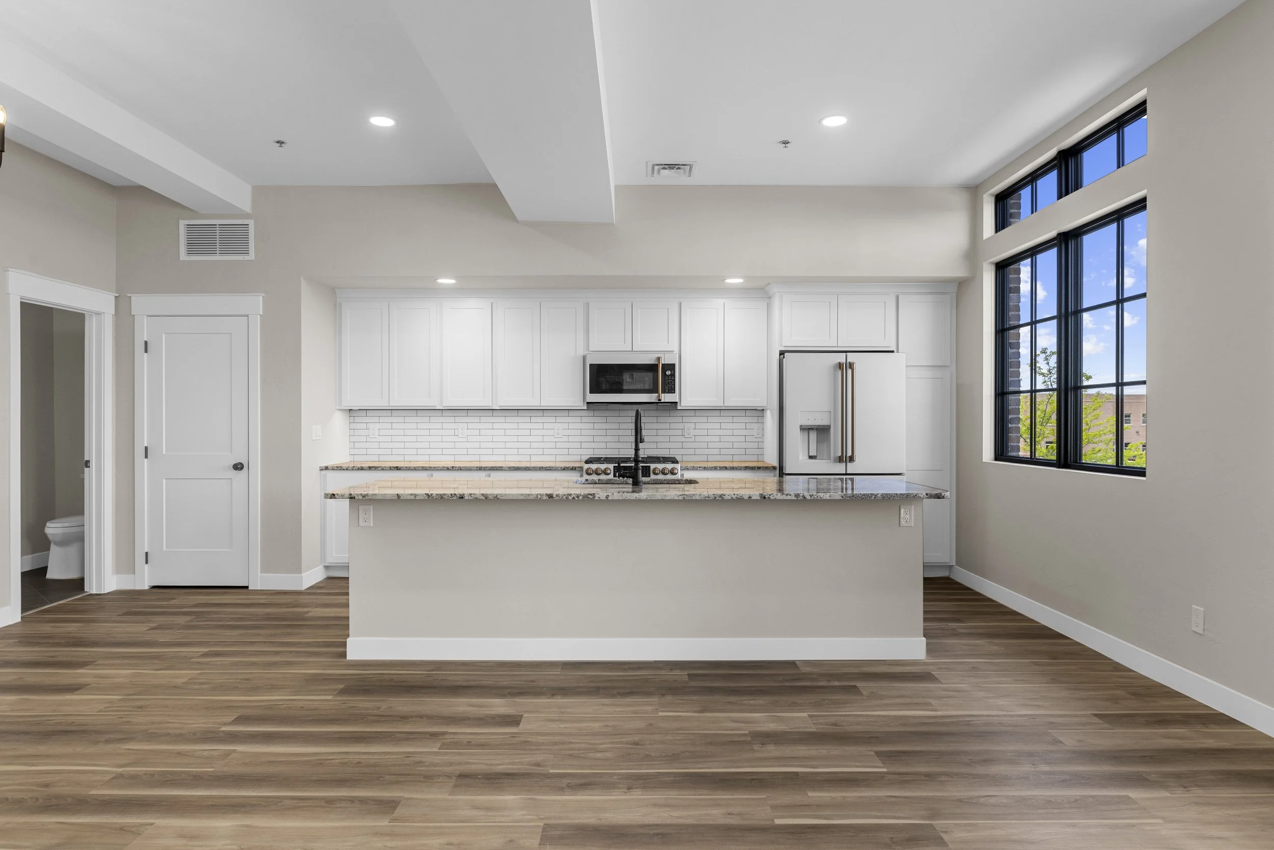 Modern kitchen with white cabinets, granite countertops, a stove, microwave, refrigerator, large window showing outside view, and hardwood floors.