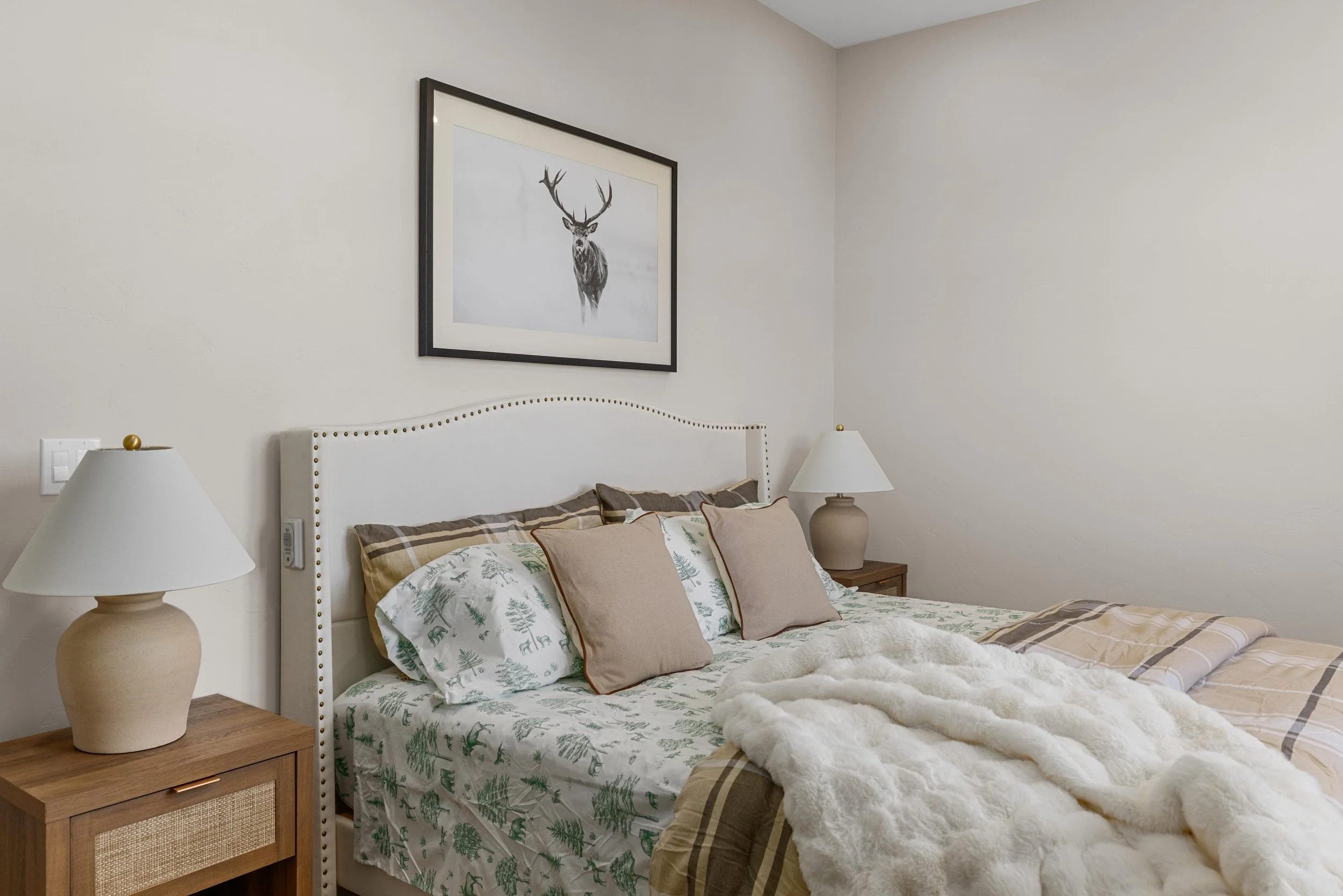 A cozy bedroom with a white headboard, earth-toned pillows, plaid bedding, and a plush white blanket. Two bedside tables each hold a beige lamp, and a framed black-and-white picture of a deer hangs above the bed.