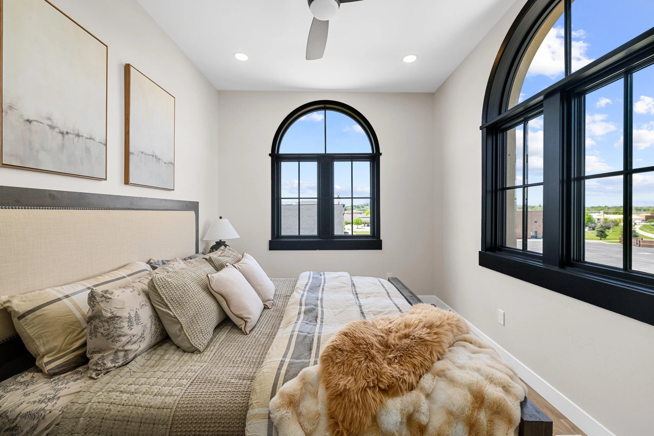 A bedroom with a large bed, beige headboard, pillows, bedside lamp, and two black-framed arched windows showing a partly cloudy sky and buildings outside.