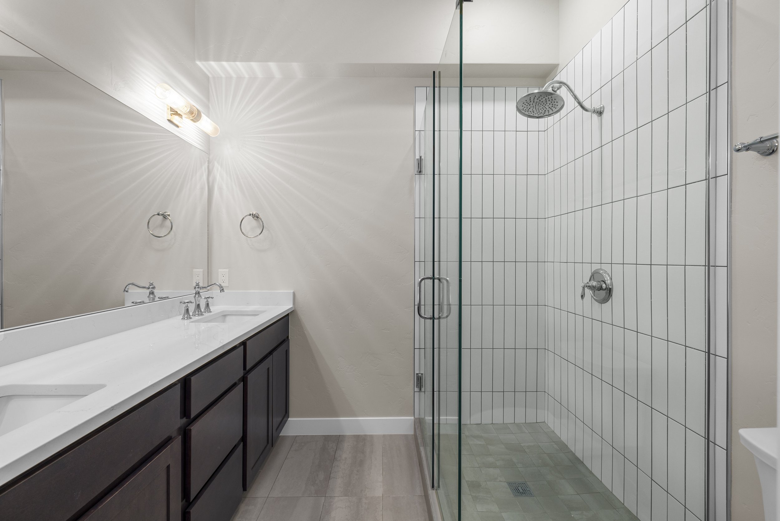 A modern bathroom with a dual-sink vanity with dark cabinets and a large mirror. To the right, there is a glass-enclosed shower with white tiled walls and a rainfall showerhead.