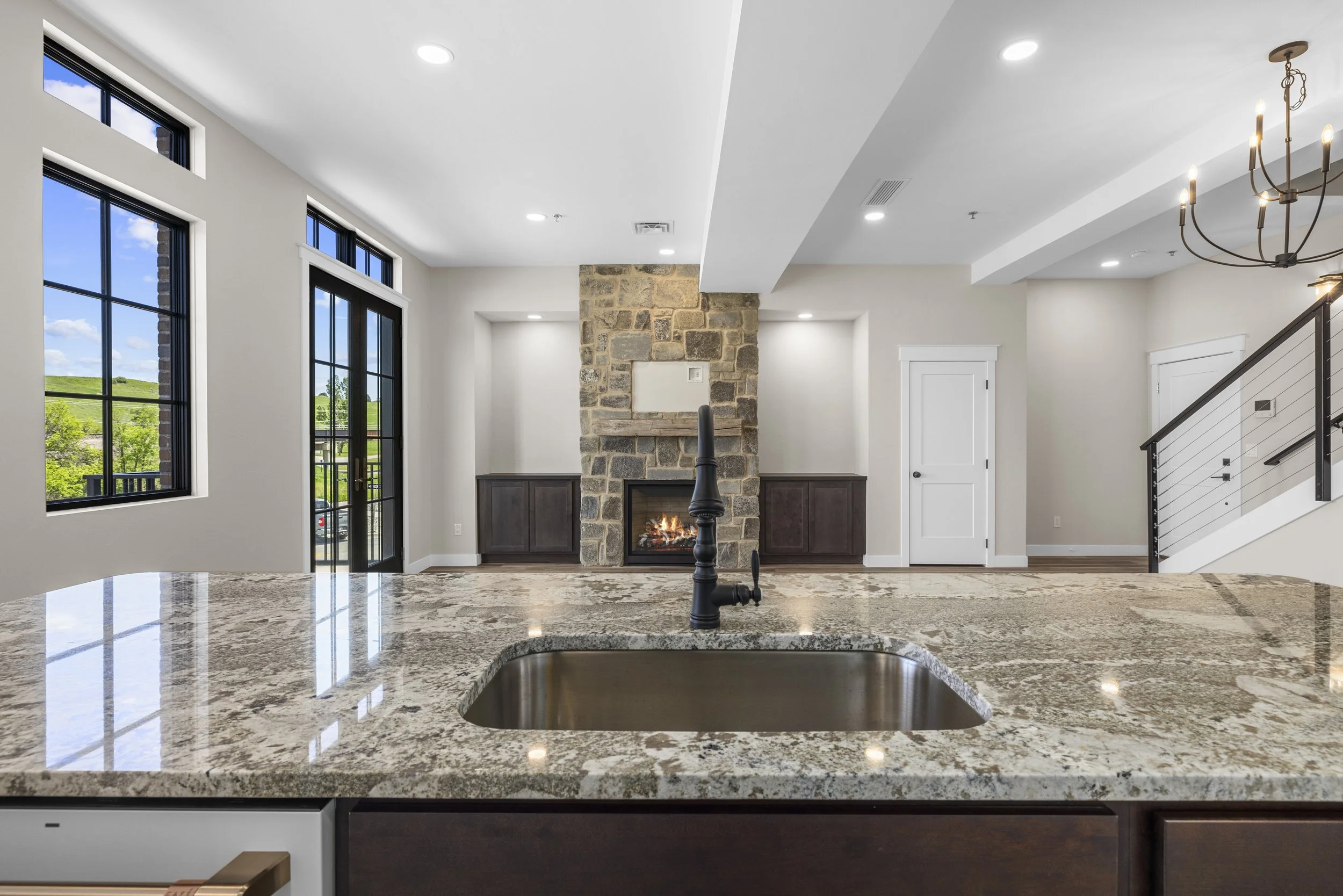 Modern kitchen with granite countertop, stainless steel sink, black faucet, leading to a spacious living room with a stone fireplace, built-in cabinets, and large windows showing greenery outside.