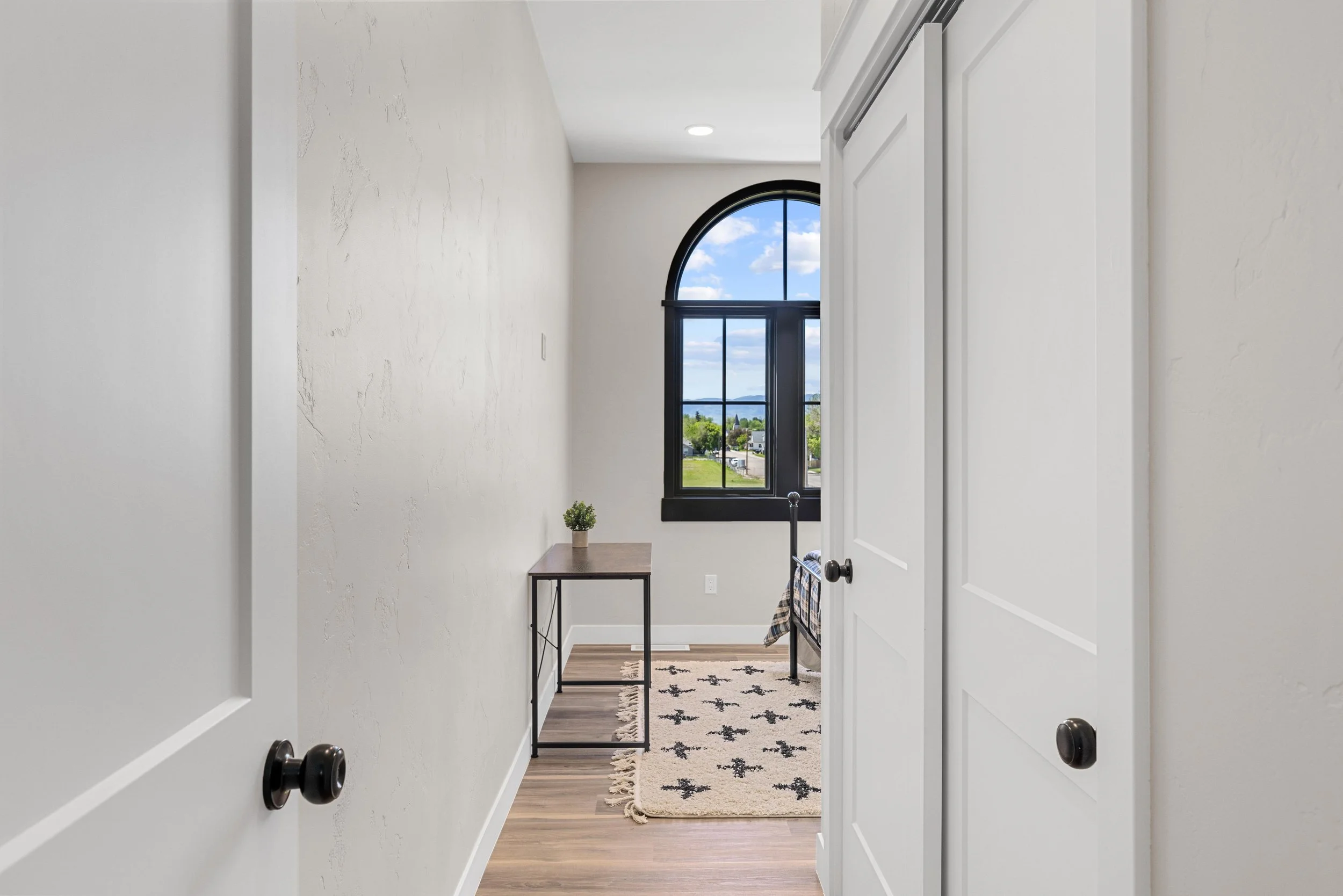 View of a bright room with a large arched window showing an outdoor scene with blue sky and trees. There is a small table with a potted plant near the window, a bed with a blanket, and a rug with a star pattern on wooden flooring.