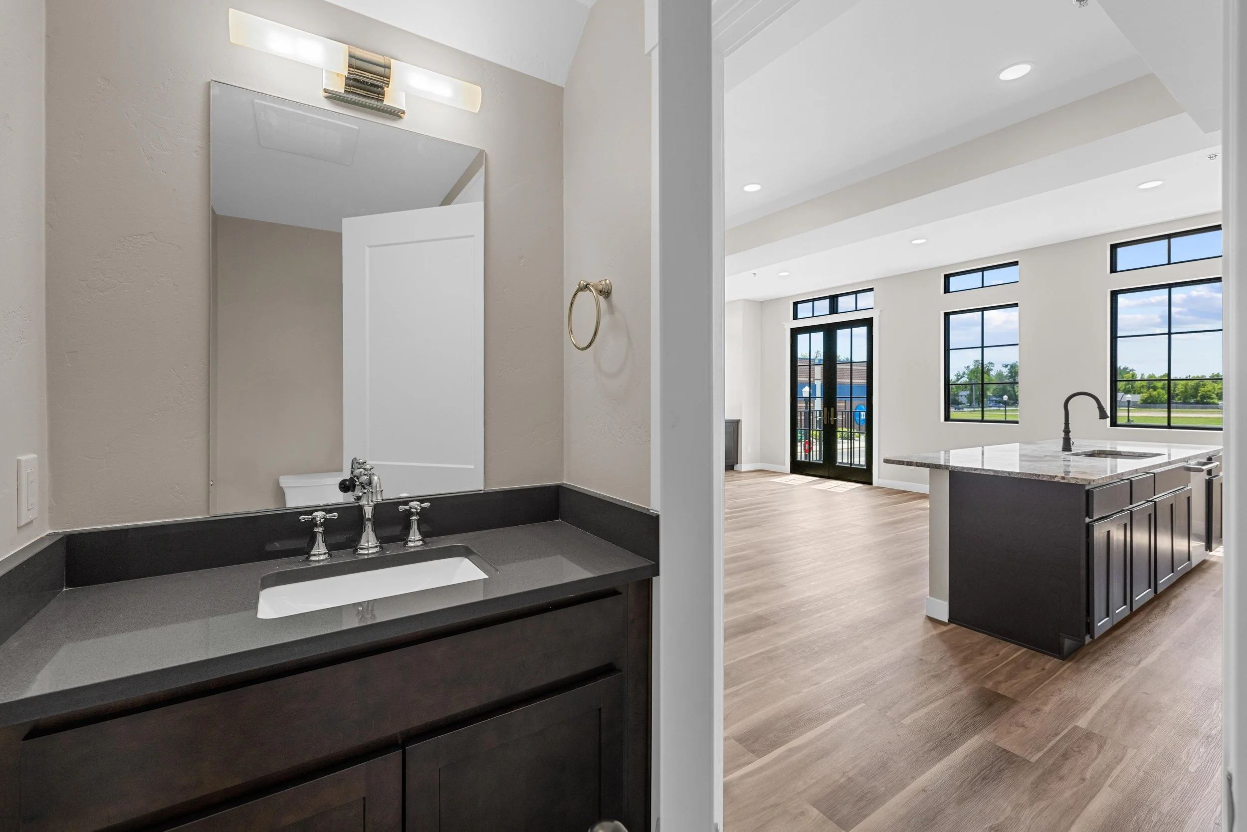 Interior view of a modern home with a bathroom vanity area on the left and a spacious kitchen and living area on the right, featuring large windows, wood flooring, and an island with a sink in the kitchen.