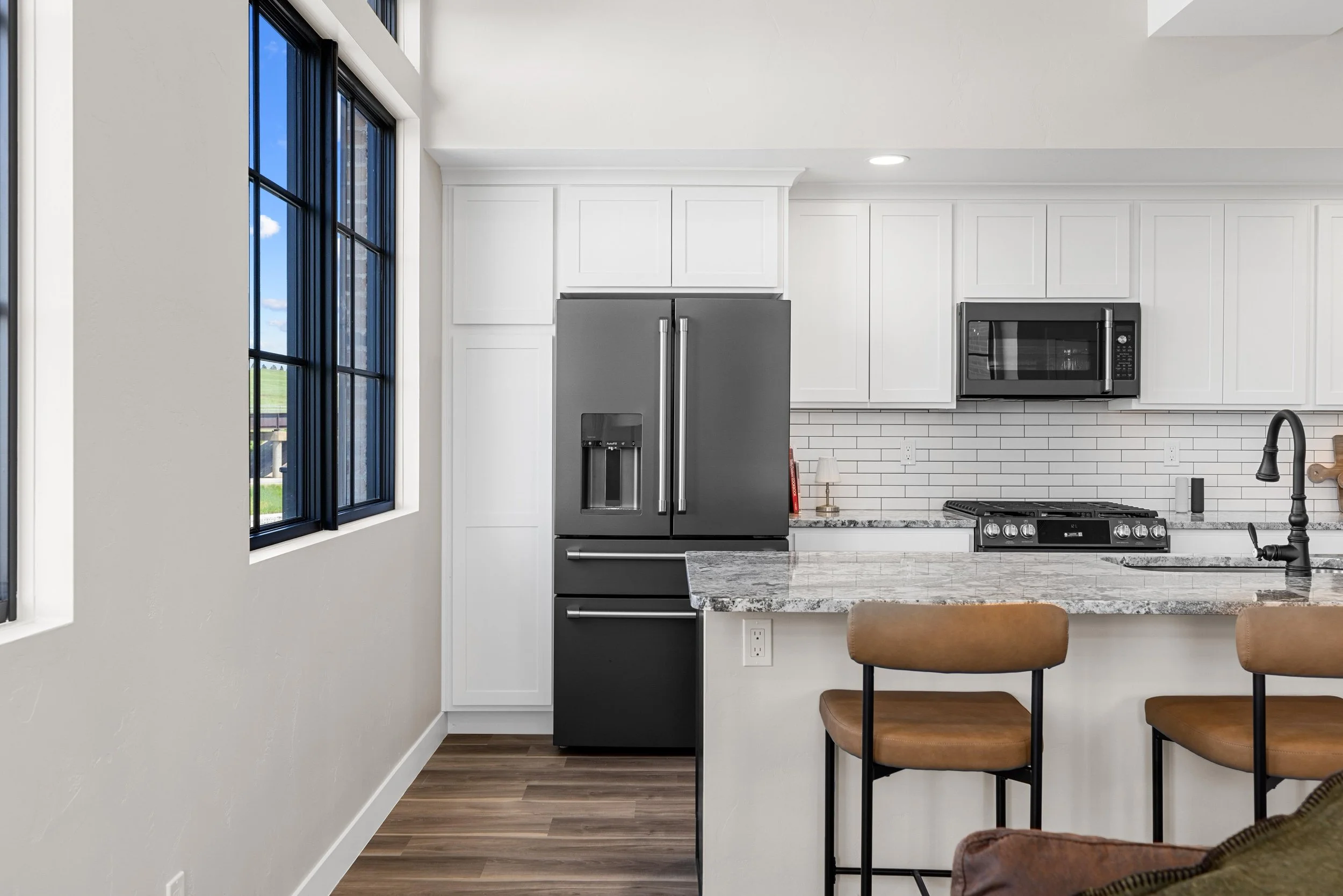 Modern kitchen with white cabinets, black appliances including a refrigerator, microwave, and stove, a marble countertop, a double sink with black faucet, and two brown barstools at the island. There are two large windows with a view outside.