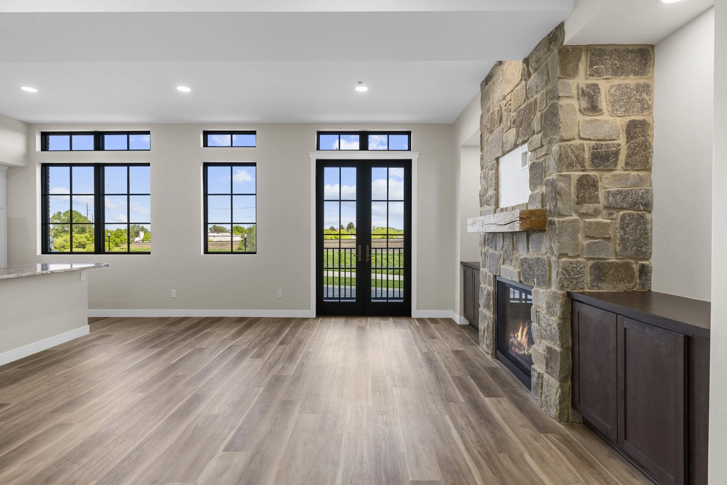 Empty living room with large windows, glass double doors, a stone fireplace with wooden mantel, and hardwood flooring.