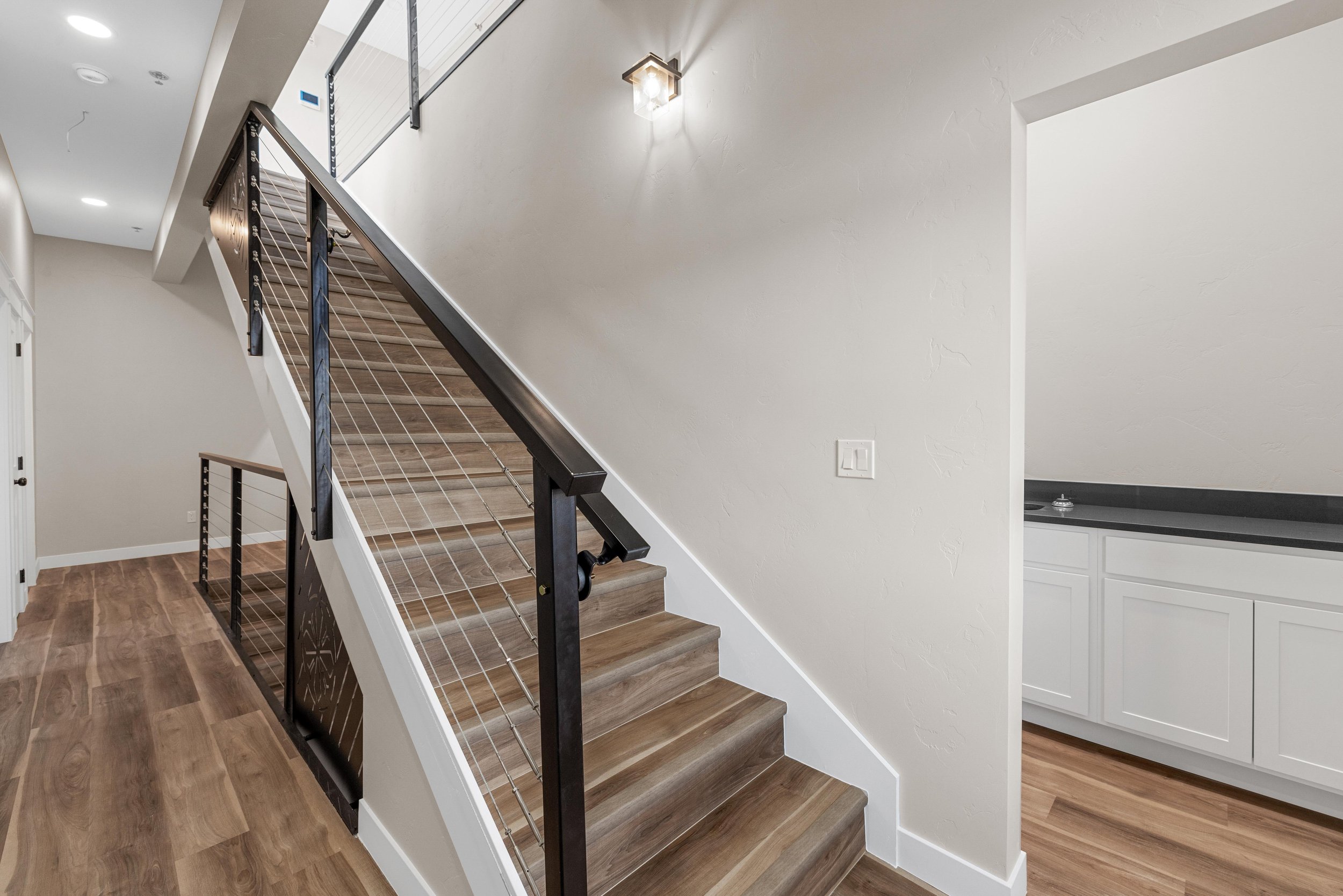 Interior view of a modern staircase with wooden steps, black metal handrail, and cable railing, adjacent to a small kitchenette with white cabinets and a black countertop.