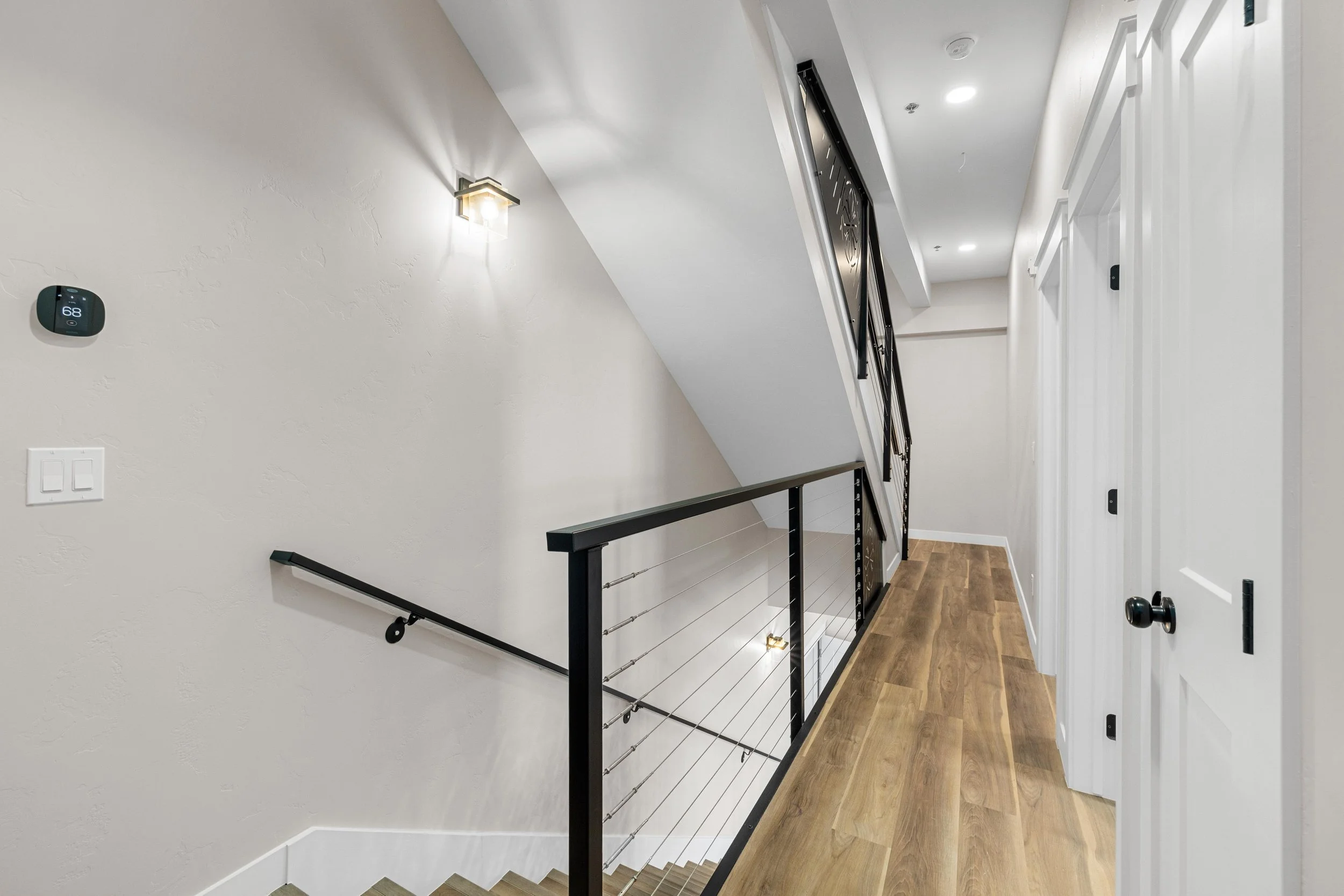 Upscale staircase landing with wood flooring, black metal railing, white textured walls, wall-mounted light fixtures, thermostat, and closed white doorsin an Ice Haus Condo for sale in Sheridan, Wyoming.