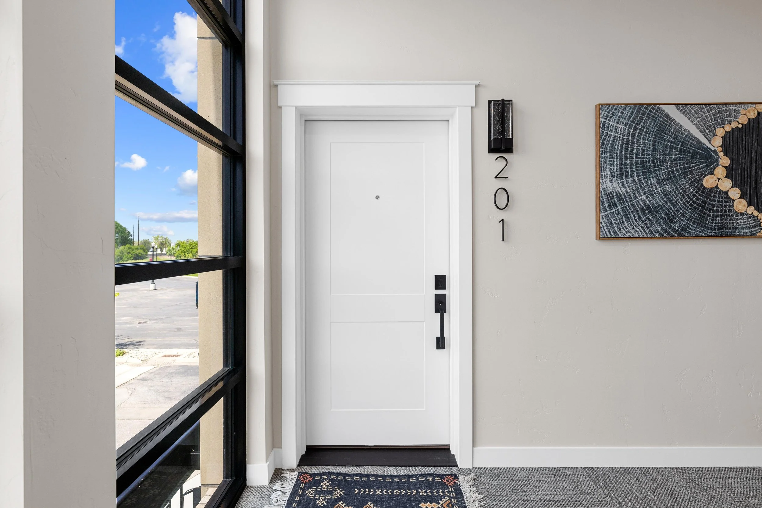 Interior of a building with a white front door, black door handle, black wall-mounted light, and a framed art piece on the wall. The door has the number '201' vertically displayed beside it. There is a large window showing an outdoor parking lot and 