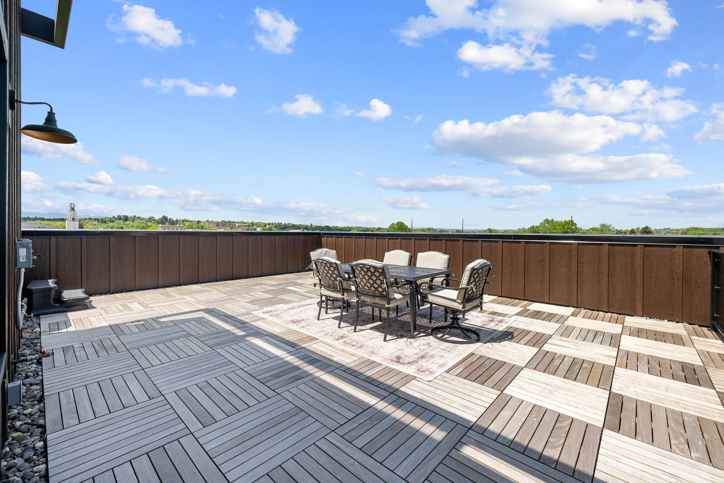 Rooftop patio with outdoor furniture, wooden and tile flooring, and a brown wooden railing, overlooking a blue sky with white clouds and green landscape.
