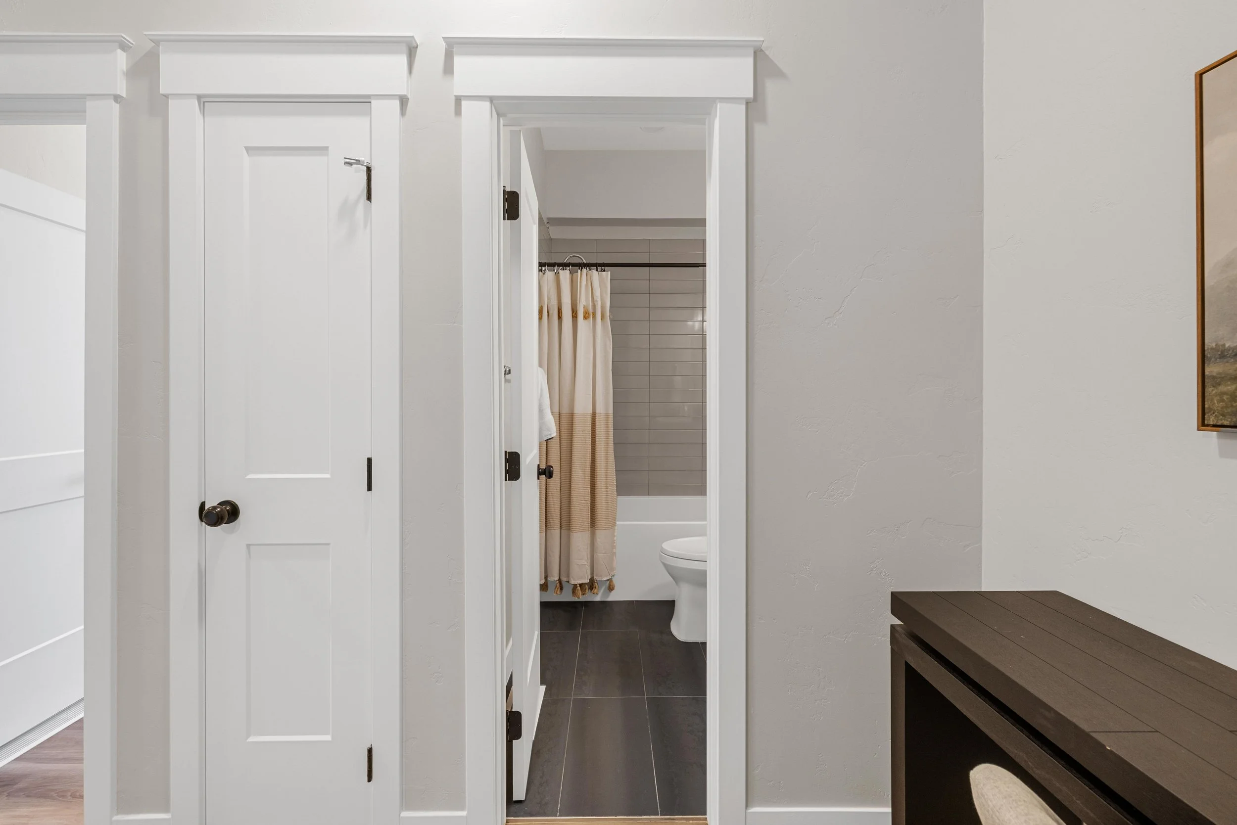 View into a bathroom through an open door, showing a toilet and a bathtub with a beige and white shower curtain. The door is in a white room with light-colored walls and dark flooring, in an Ice Haus Condo for sale in Sheridan, Wyoming.