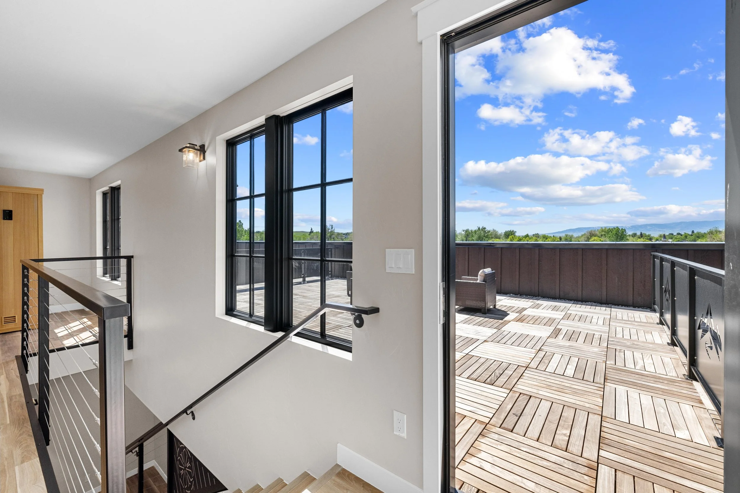 Interior view of a modern home with a staircase and large black-framed windows, opening to a spacious outdoor wooden deck with seating, overlooking a scenic landscape with a mountain range and partly cloudy sky.