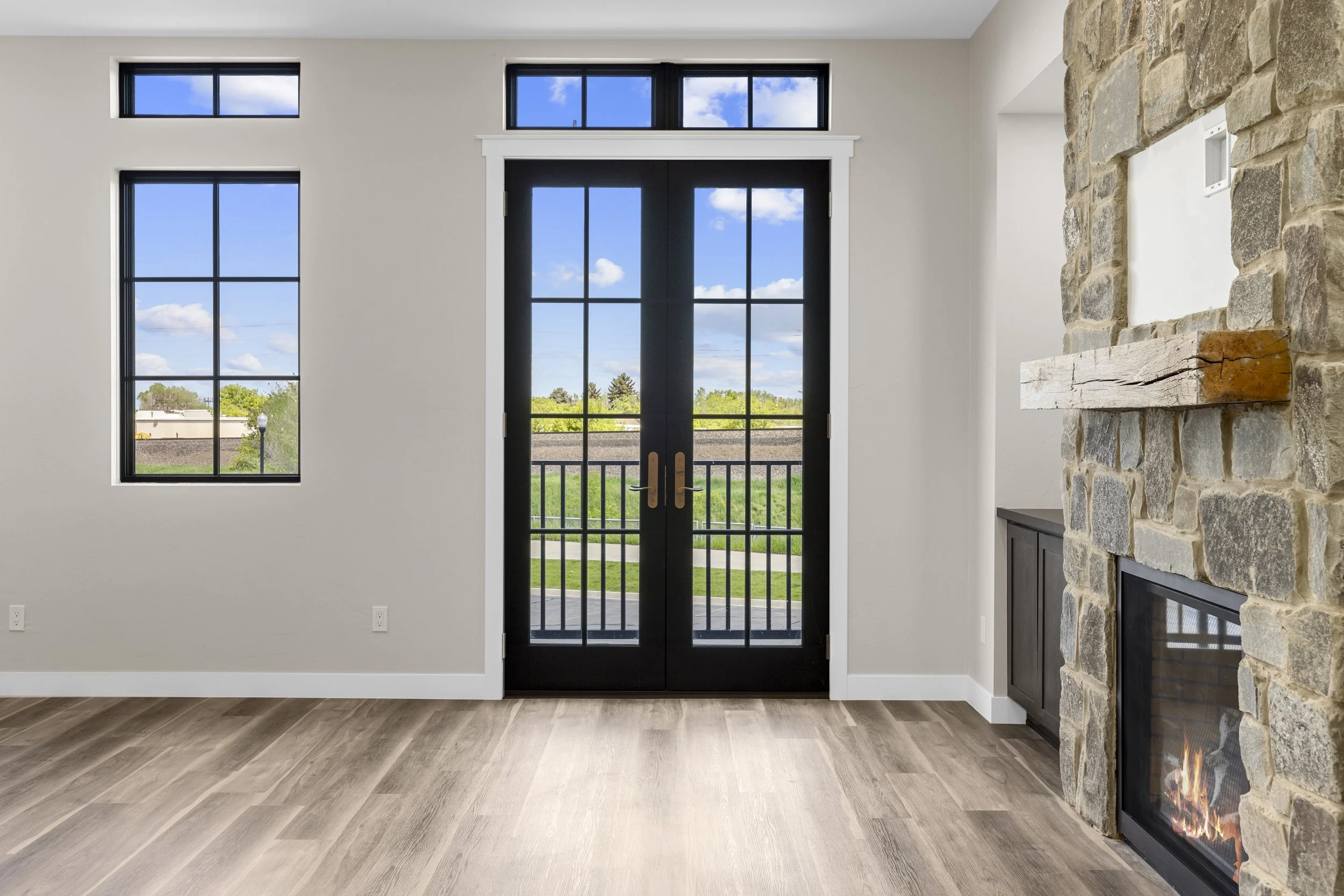 Living room with a stone fireplace, black double doors with glass panes, and three large windows showing a green outdoor area under a blue sky with clouds.