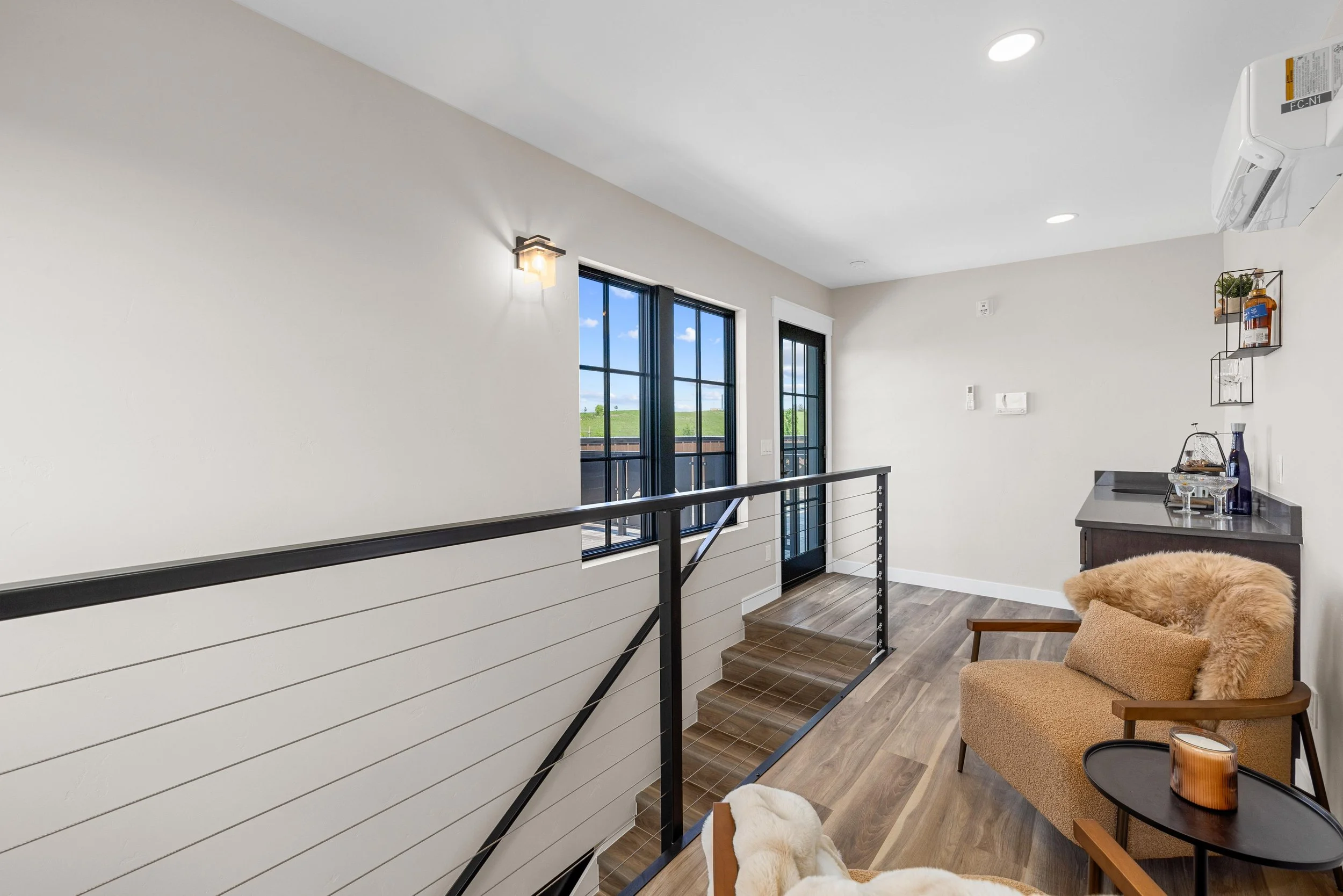 Interior of a modern upstairs landing area with three black-framed windows, a glass door, a beige armchair with a matching pillow, a small black side table with a candle, and a wooden cabinet with decorative items on top.