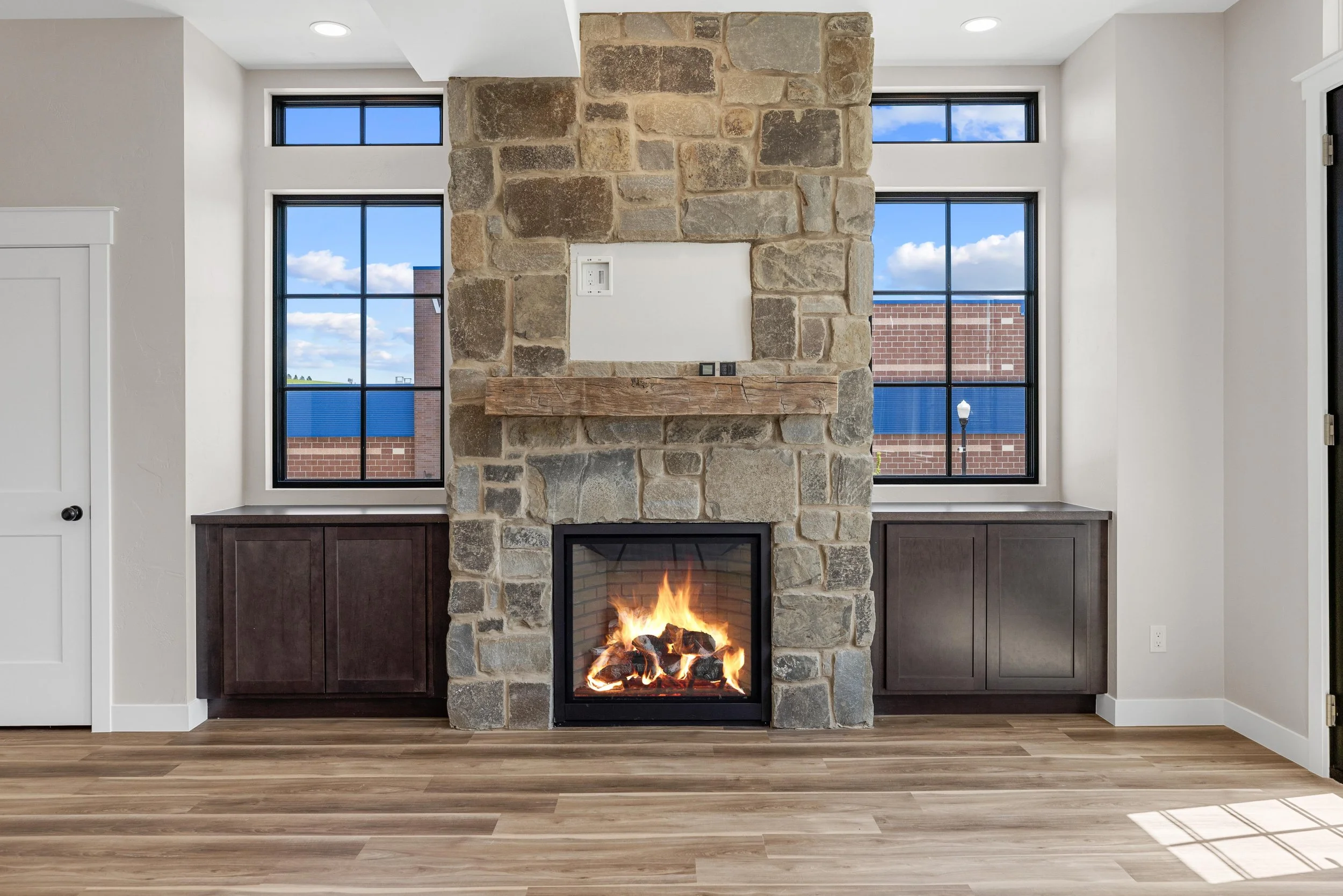 Living room in an Ice Haus Condo for sale in Sheridan, Wyoming, with a stone fireplace, two large windows on either side, dark wood cabinets below the windows, and a hardwood floor.