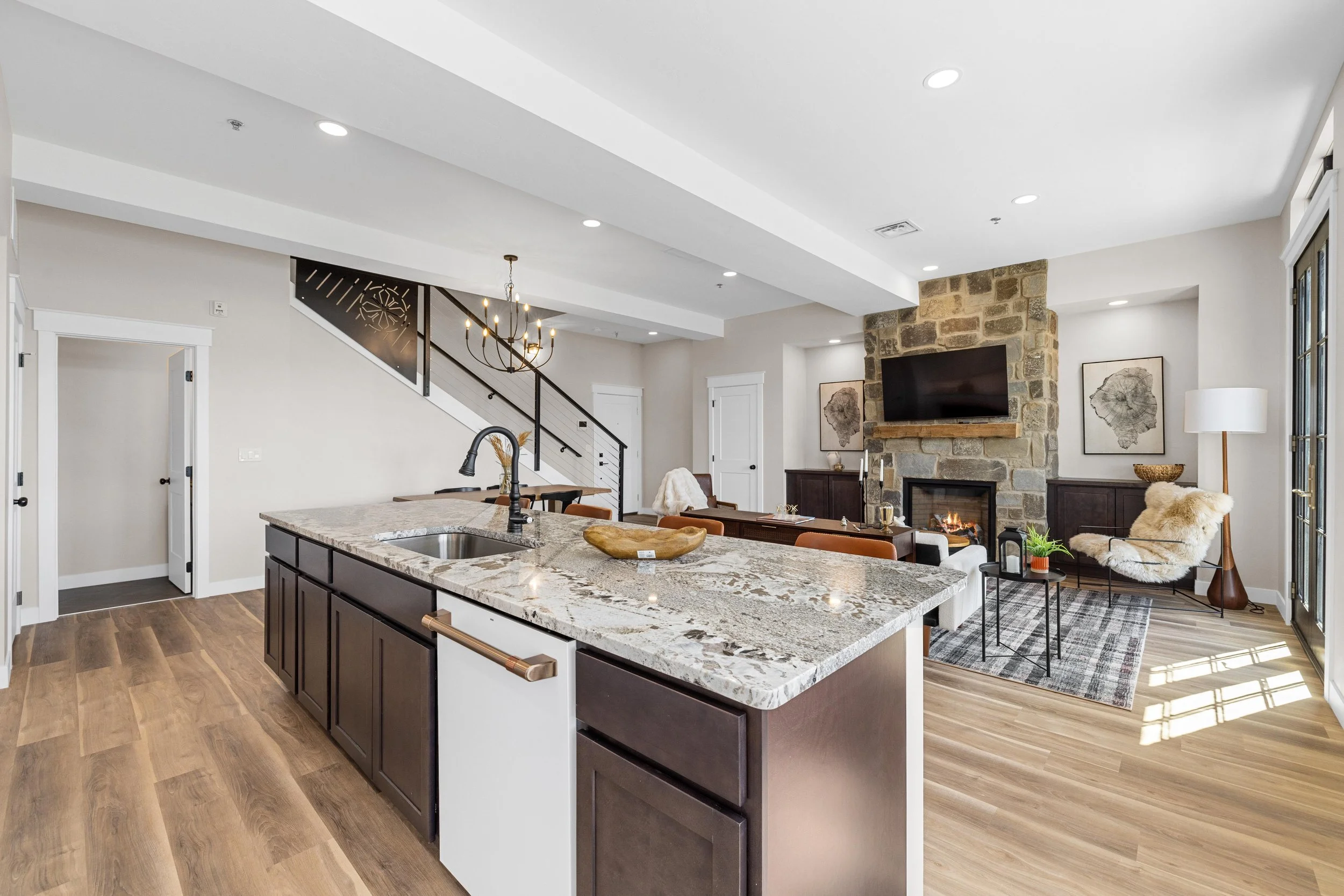 Open-concept living room with kitchen island, stone fireplace, and seating area with chairs, coffee table, and TV, illuminated by natural light from glass doors.