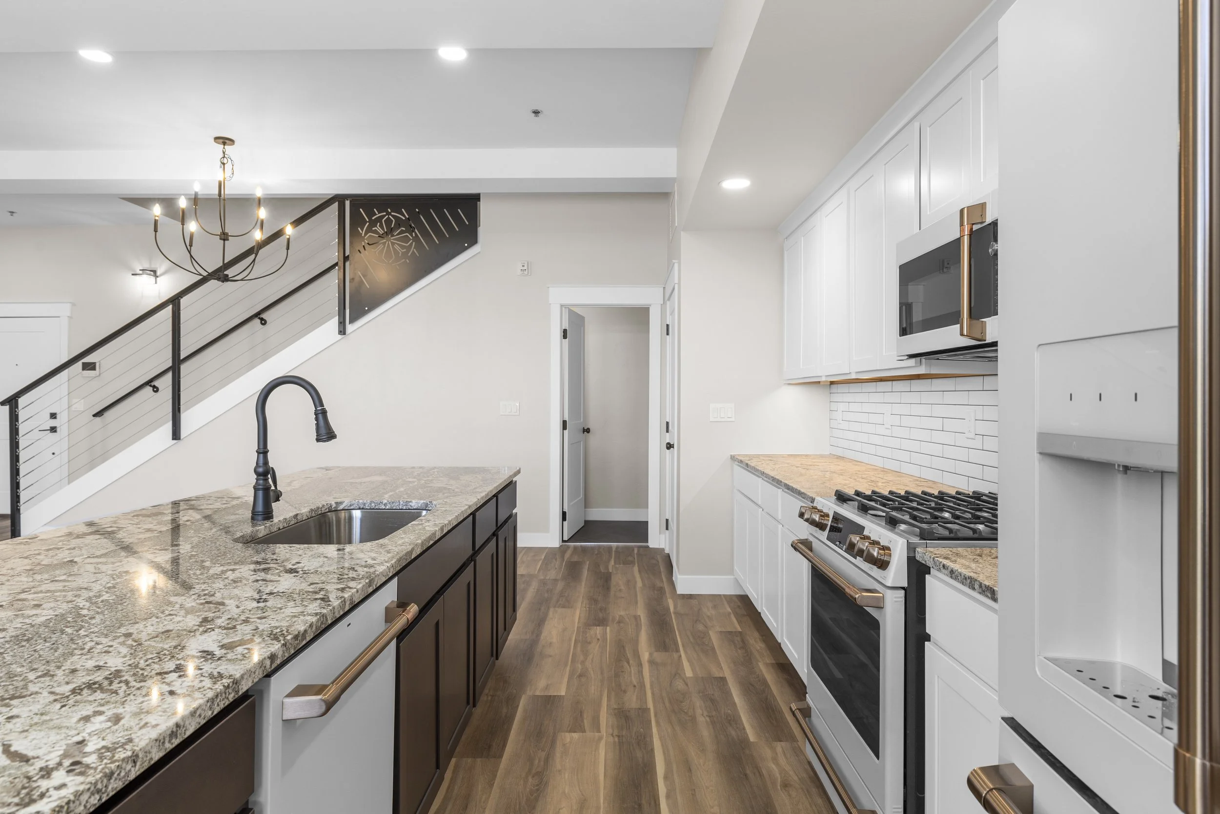 Modern kitchen with granite countertops, white cabinets, black fixtures, and hardwood flooring, adjacent to a staircase.