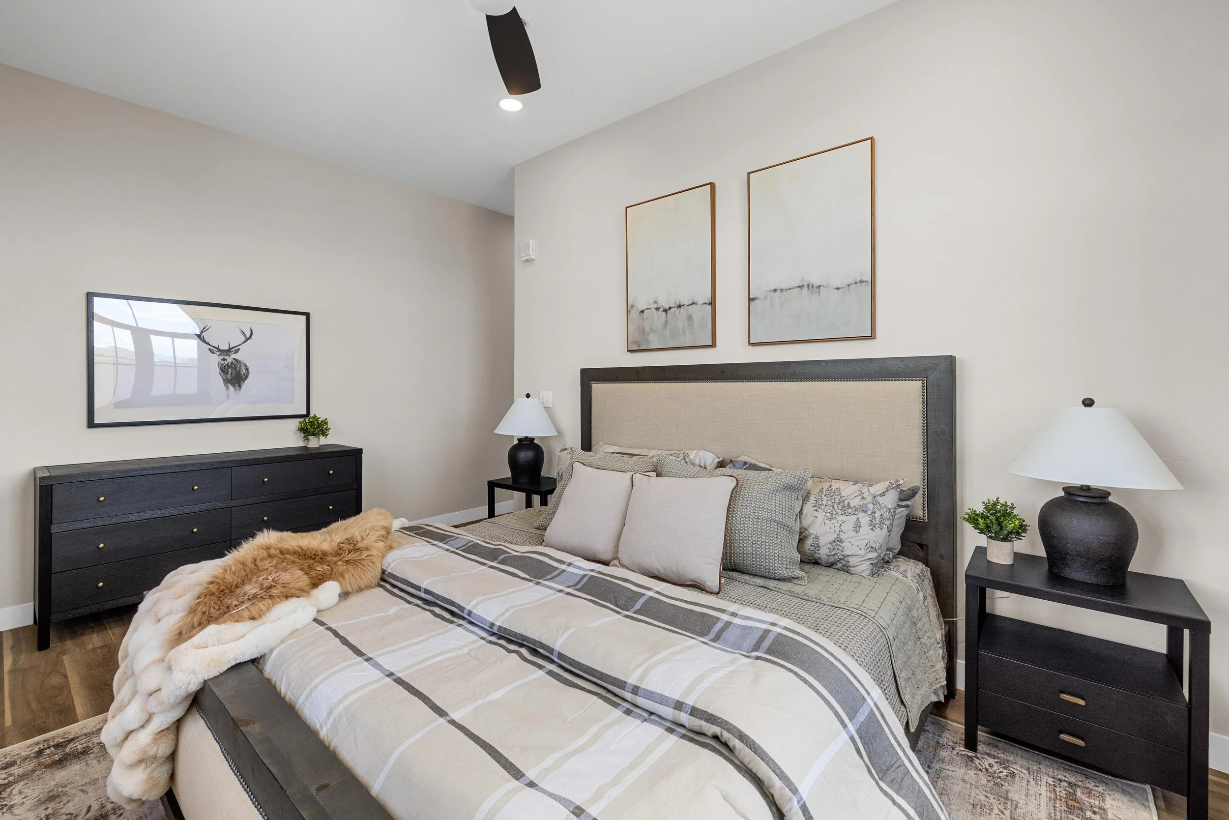 A bedroom with a large bed that has a beige headboard, multiple pillows, and a beige, gray, and white plaid comforter. There are two black nightstands with white lamps, a black dresser with brass knobs, and wall art including two abstract paintings a