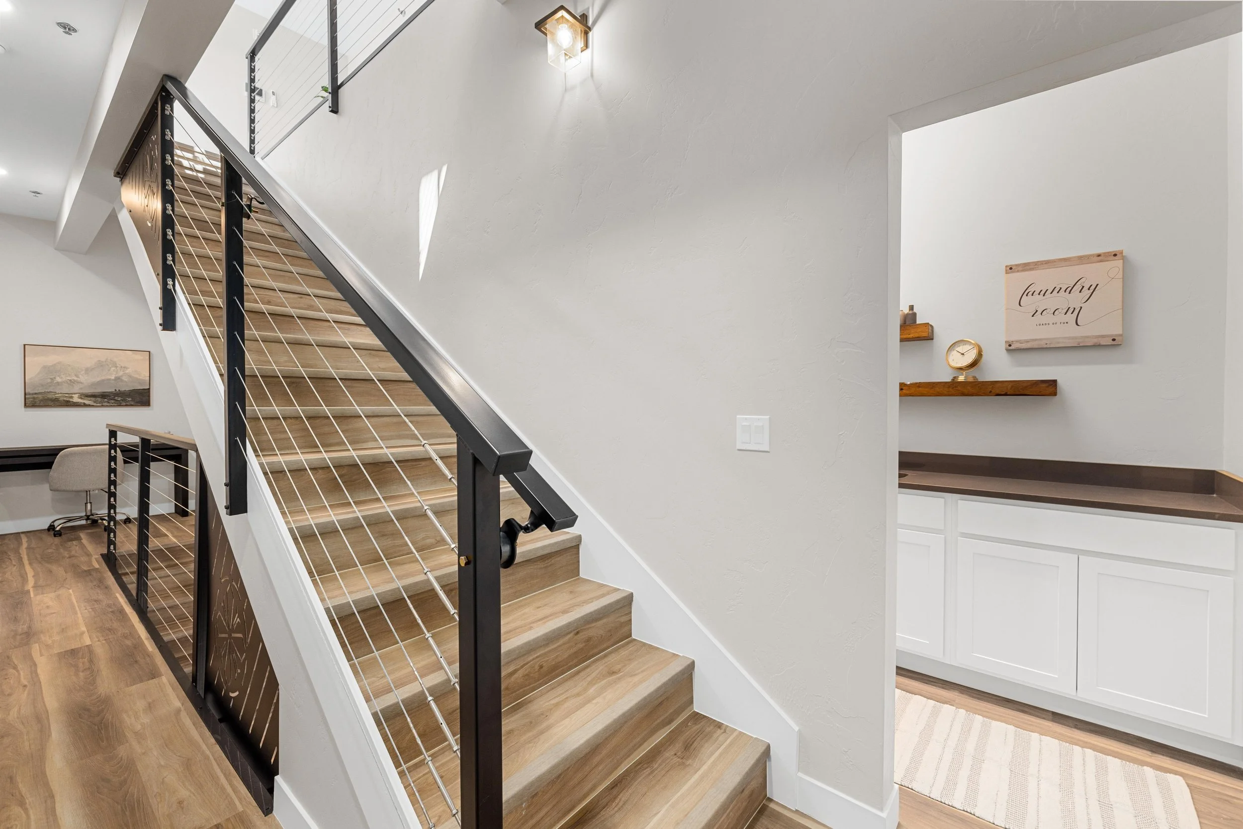 Interior view of a staircase with wooden steps, black metal railing, and white walls. There is a wall-mounted light fixture and a doorway leading to a laundry room with white cabinetry and wooden shelves.