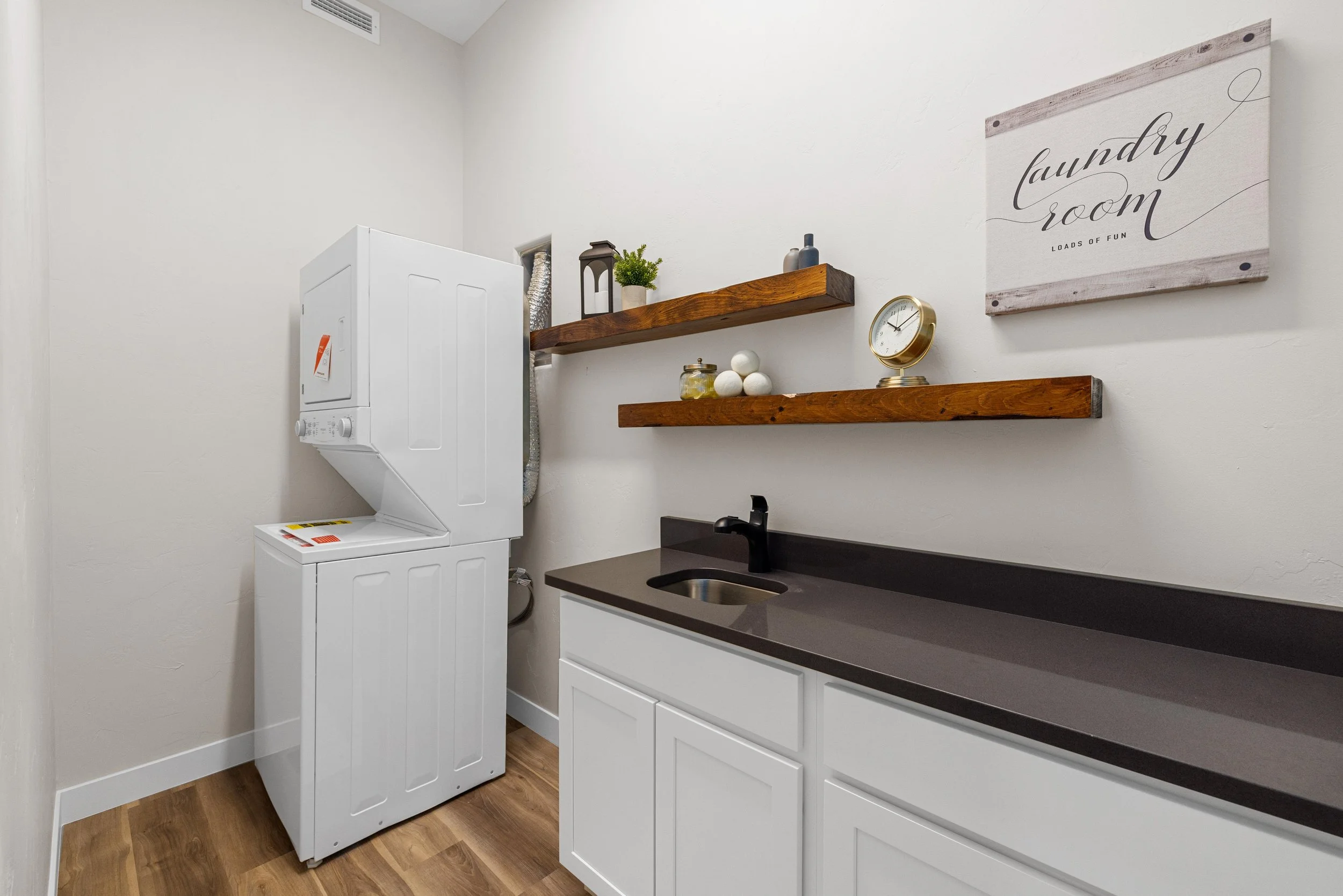 A laundry room with a stacked washer and dryer, white cabinets, a black faucet on a dark countertop, and wooden shelves with decorative items and a wall sign.