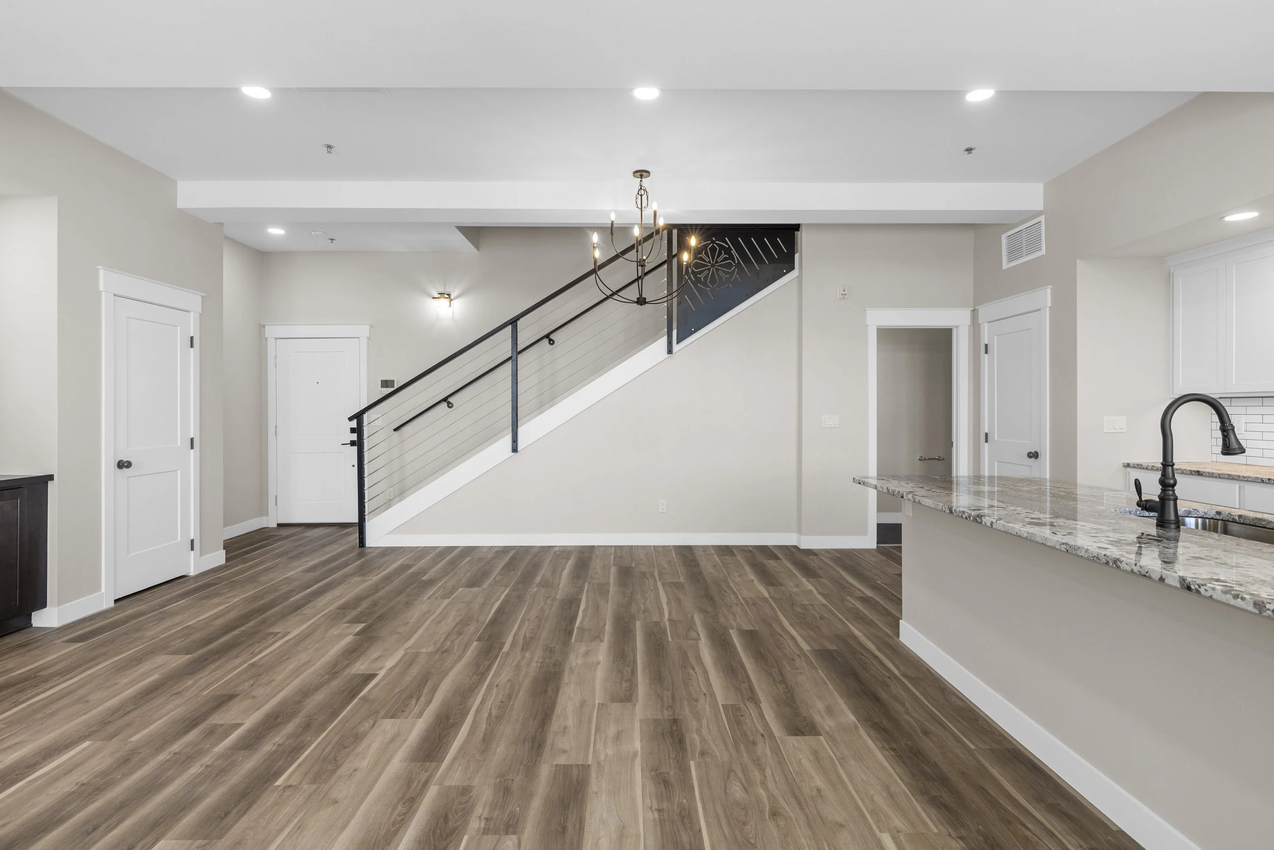 Empty living room with hardwood floors, white walls, staircase with black railing, chandelier, kitchen counter with granite top, modern appliances, in an apartment or condo.