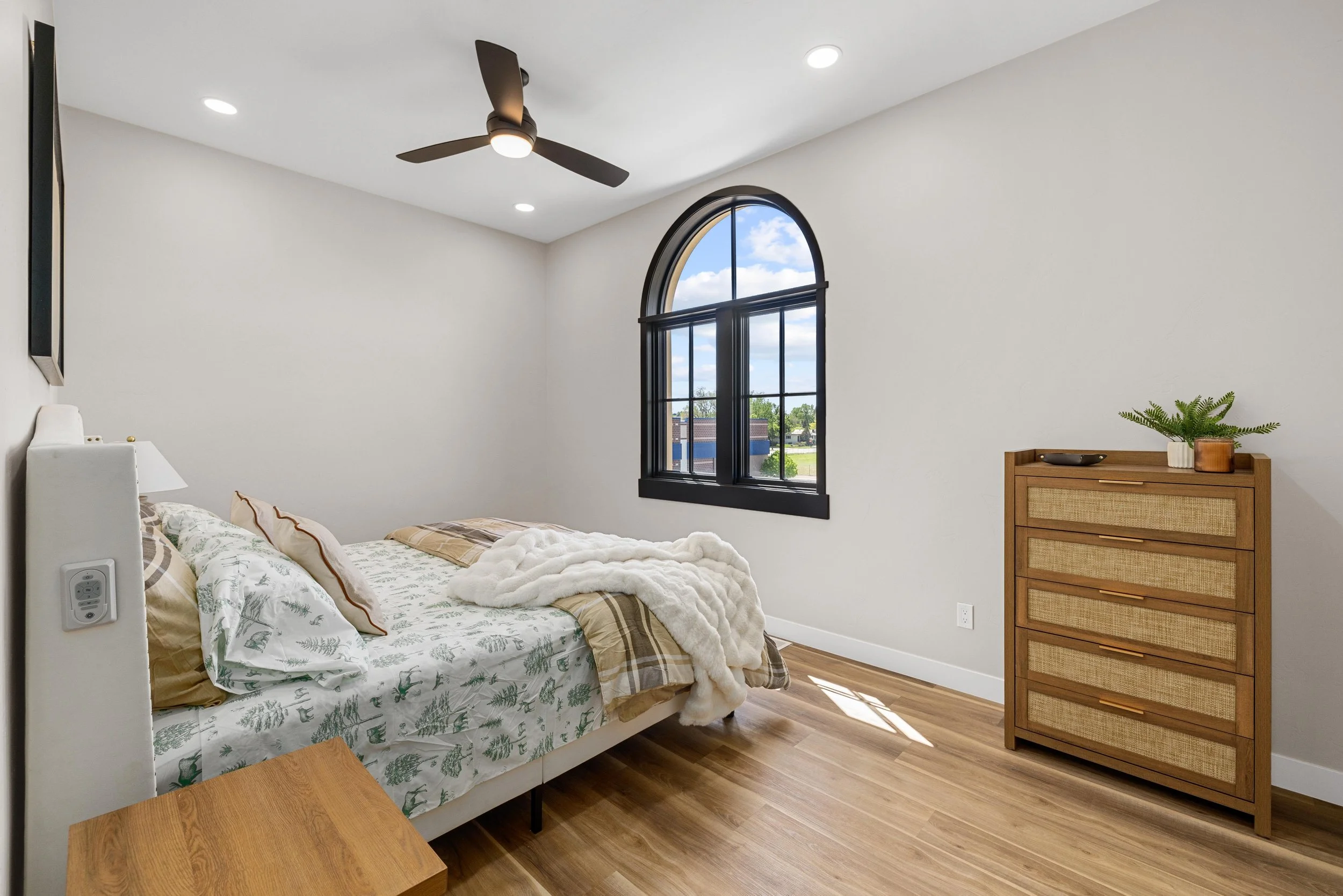 A bedroom with a white bed featuring green botanical sheets and plaid pillows, a wooden dresser with a plant on top, a black arched window showing a blue sky with clouds, and a ceiling fan.