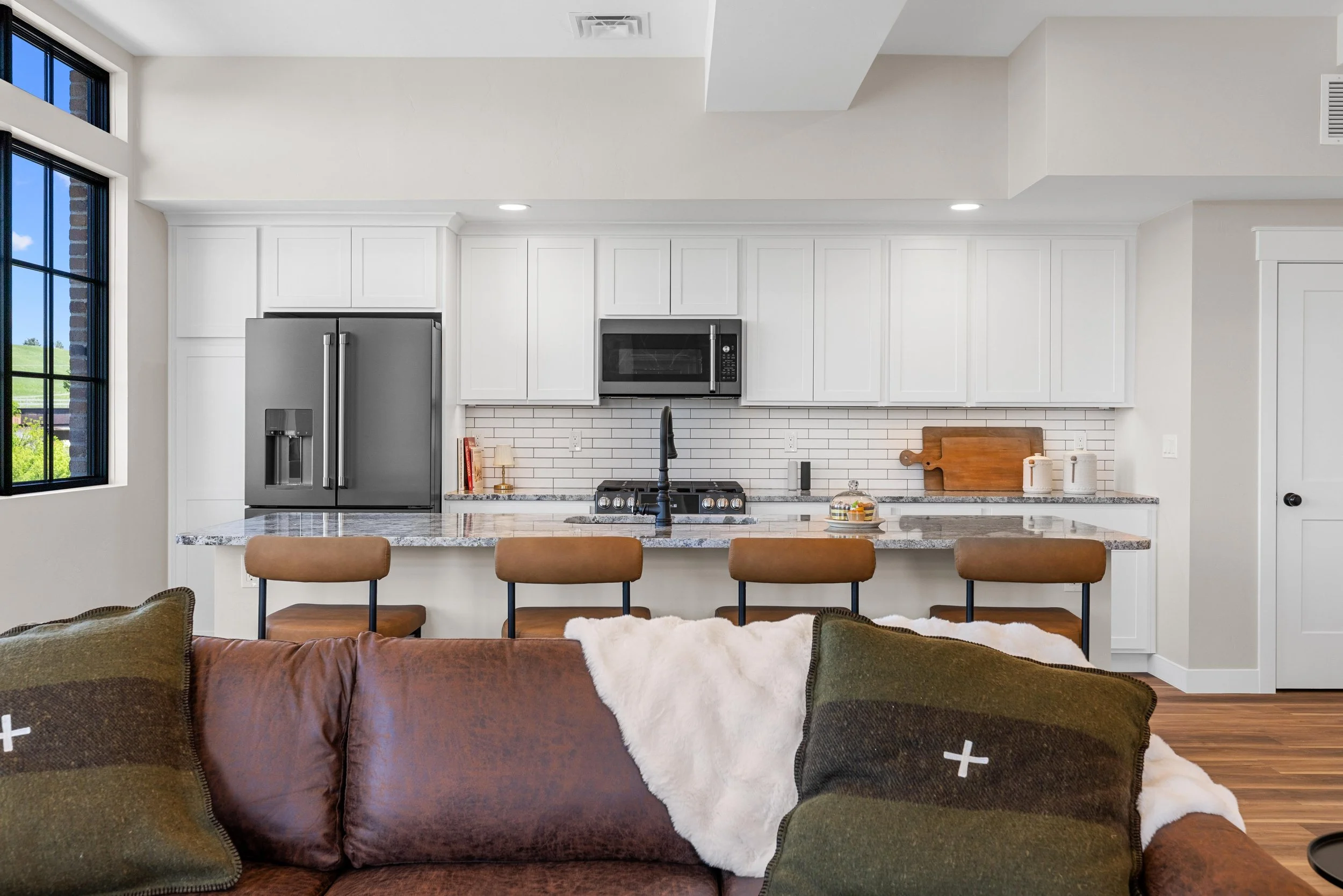 Modern kitchen with white cabinets, stainless steel refrigerator, microwave, granite countertops, and a kitchen island with seating. Living room with a brown sofa, green pillows, and a white throw blanket visible in the foreground.