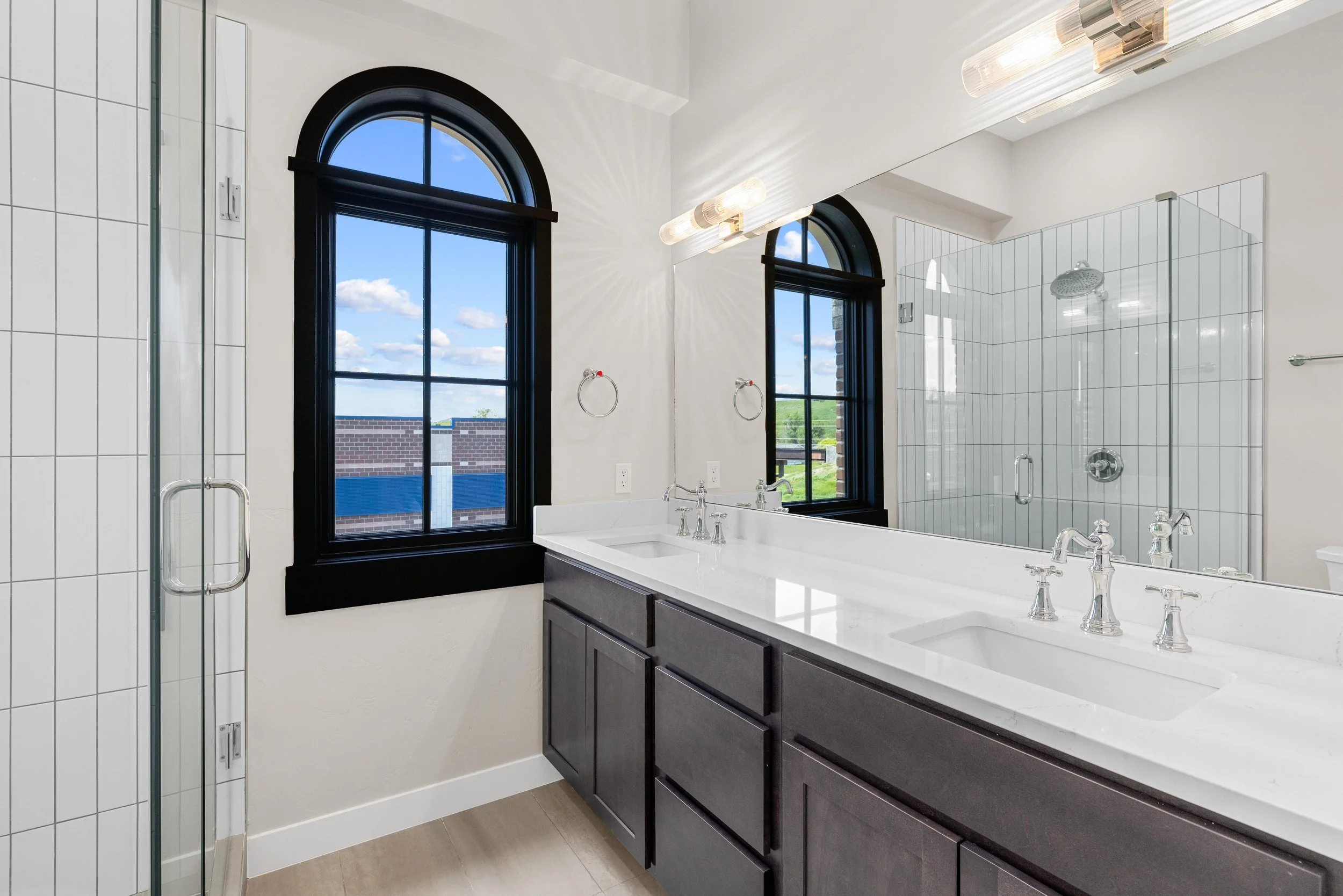 Modern bathroom with a double sink vanity, a large mirror, black framed arched windows, a glass shower enclosure with a rainfall shower head, and neutral tile flooring.