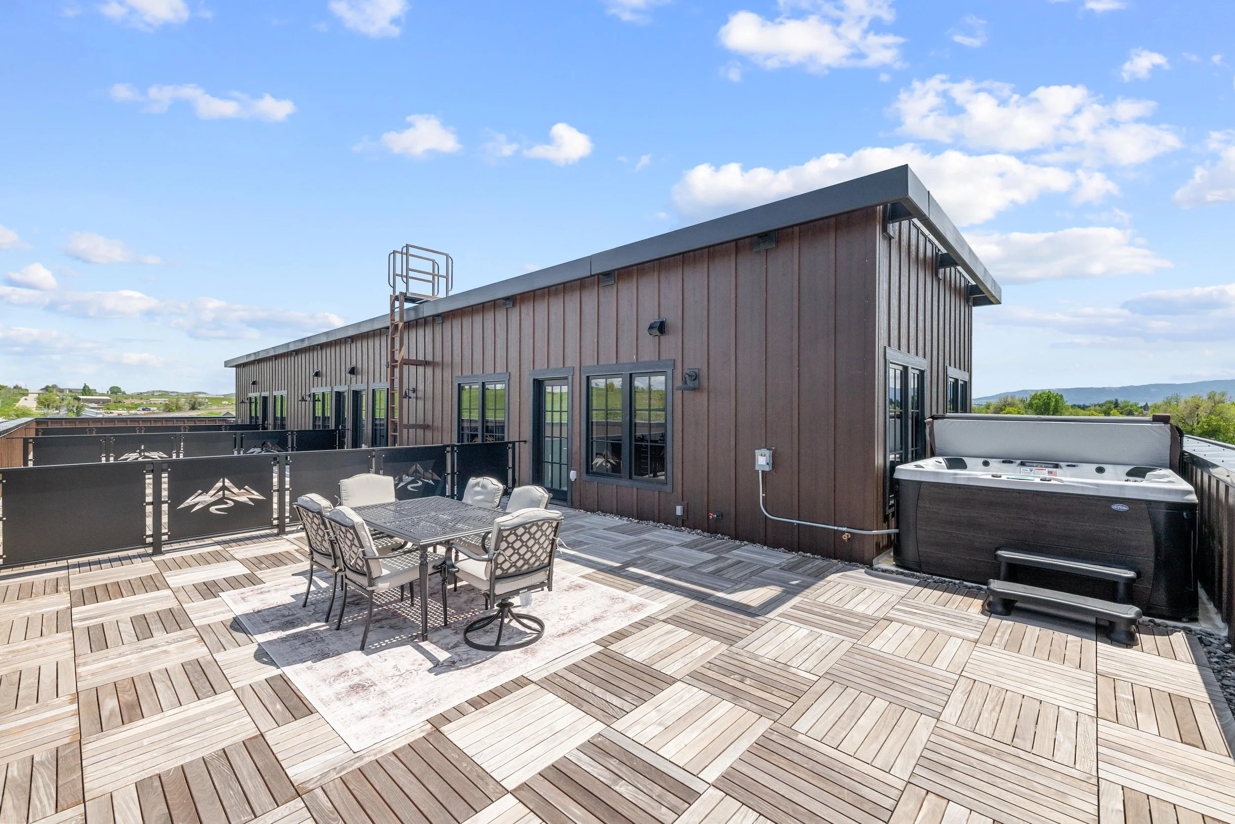 Outdoor rooftop terrace with a dining table and chairs, hot tub, and wooden decking, overlooking a rural landscape under a partly cloudy sky.