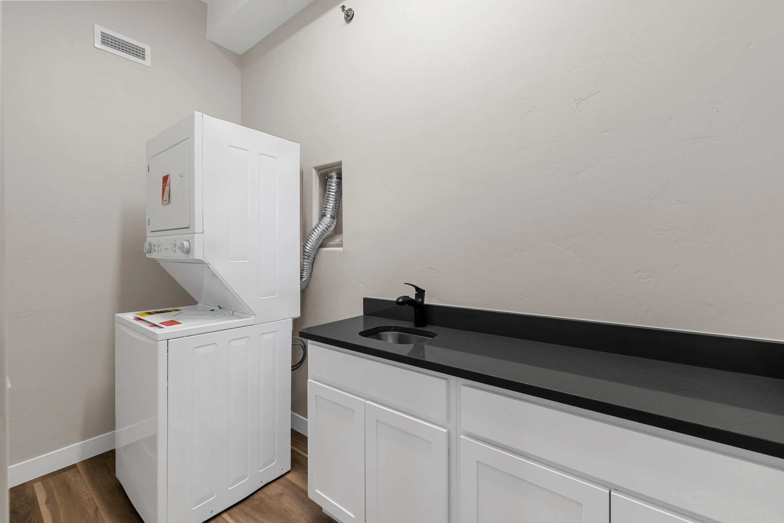 Laundry room with stacked washer and dryer, black countertop with sink, and white cabinets, hardwood floors, off-white walls, and exhaust vent.