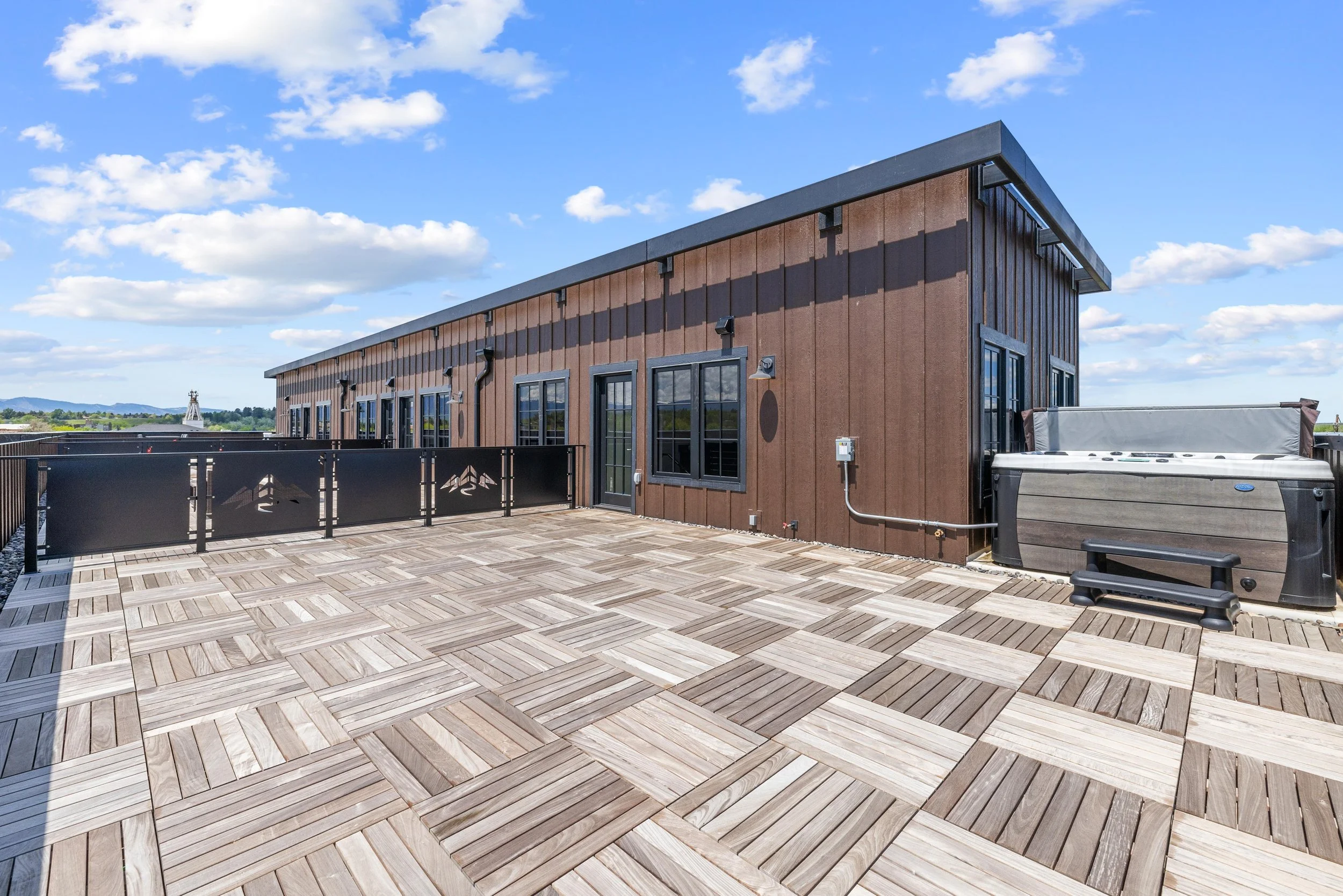 Rooftop terrace with wooden flooring, brown building wall, black railing, and hot tub under a blue sky with clouds.