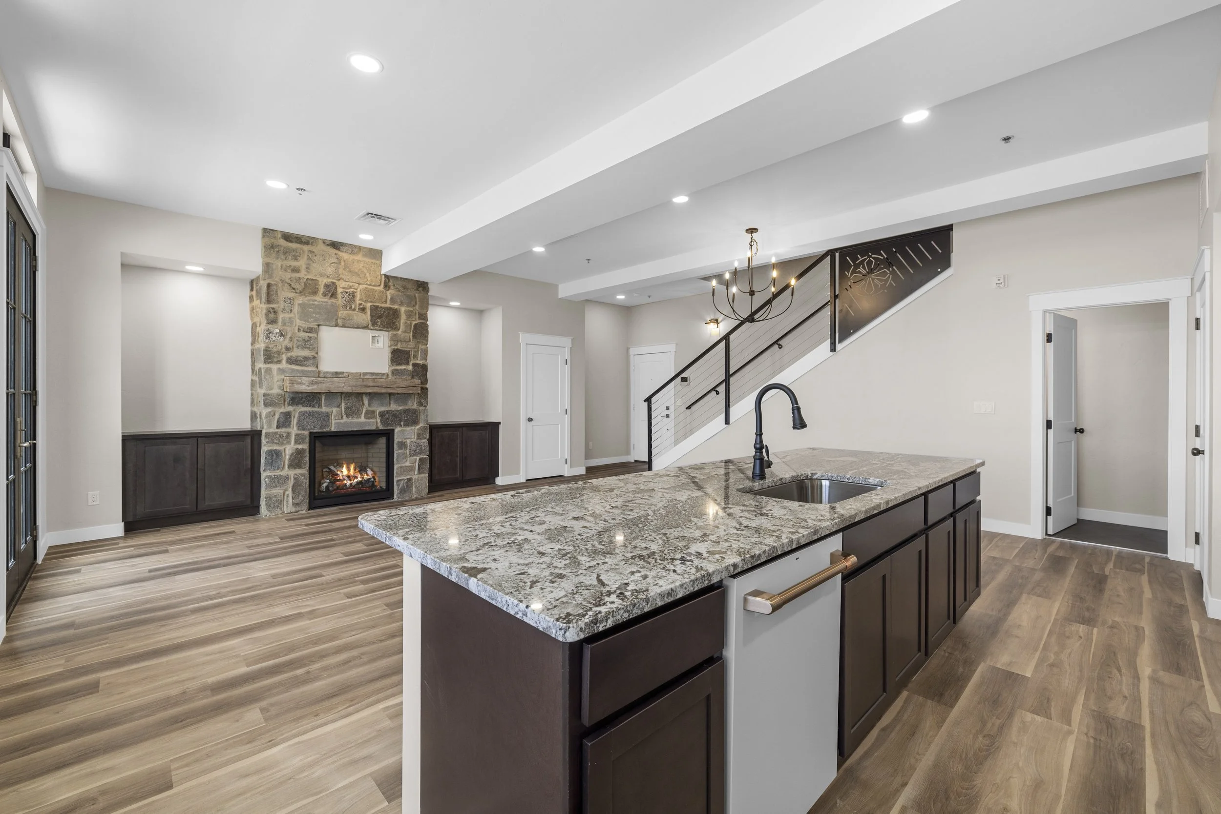 Modern open-plan kitchen and living room with a stone fireplace, wooden flooring, and a staircase with a chandelier overhead.