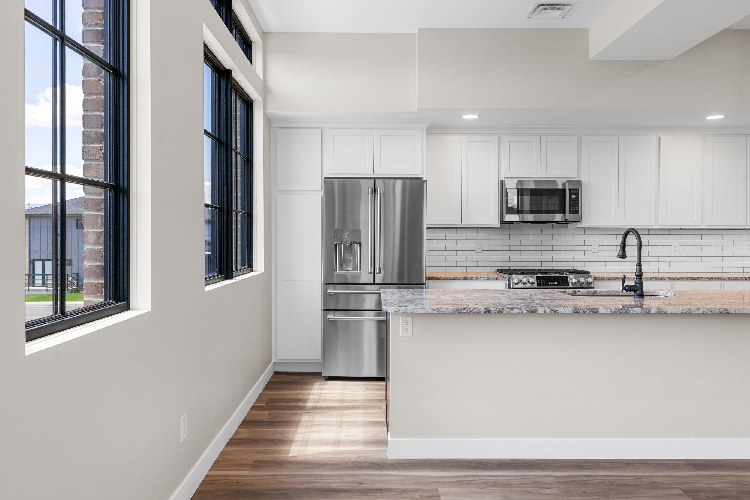 Modern kitchen with white cabinets, stainless steel refrigerator, microwave, stove, and a granite countertop island, with large windows on the left and a brick and white tile backsplash.