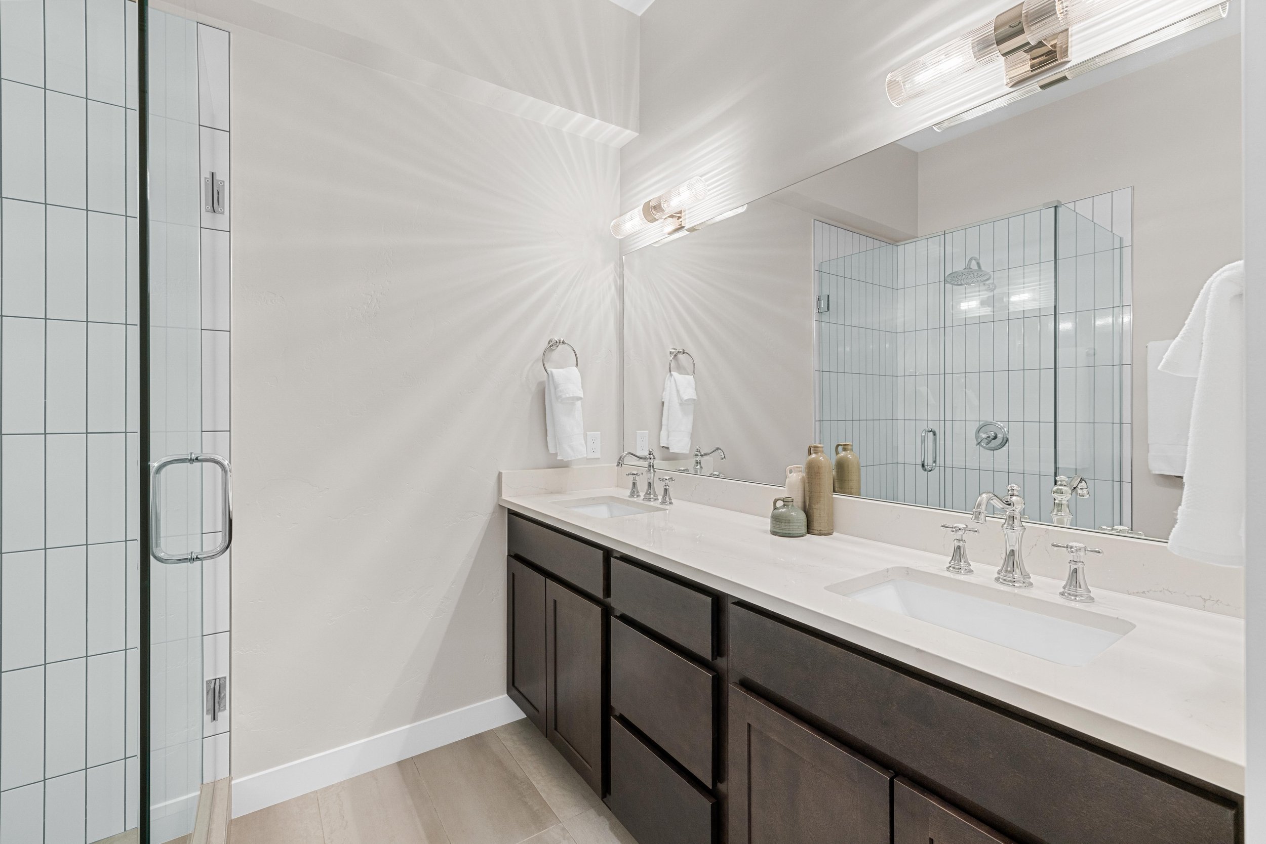 Modern bathroom with double vanity, large mirror, towel rings with white towels, decorative bottles on countertop, and a glass-enclosed shower with white tiled walls.