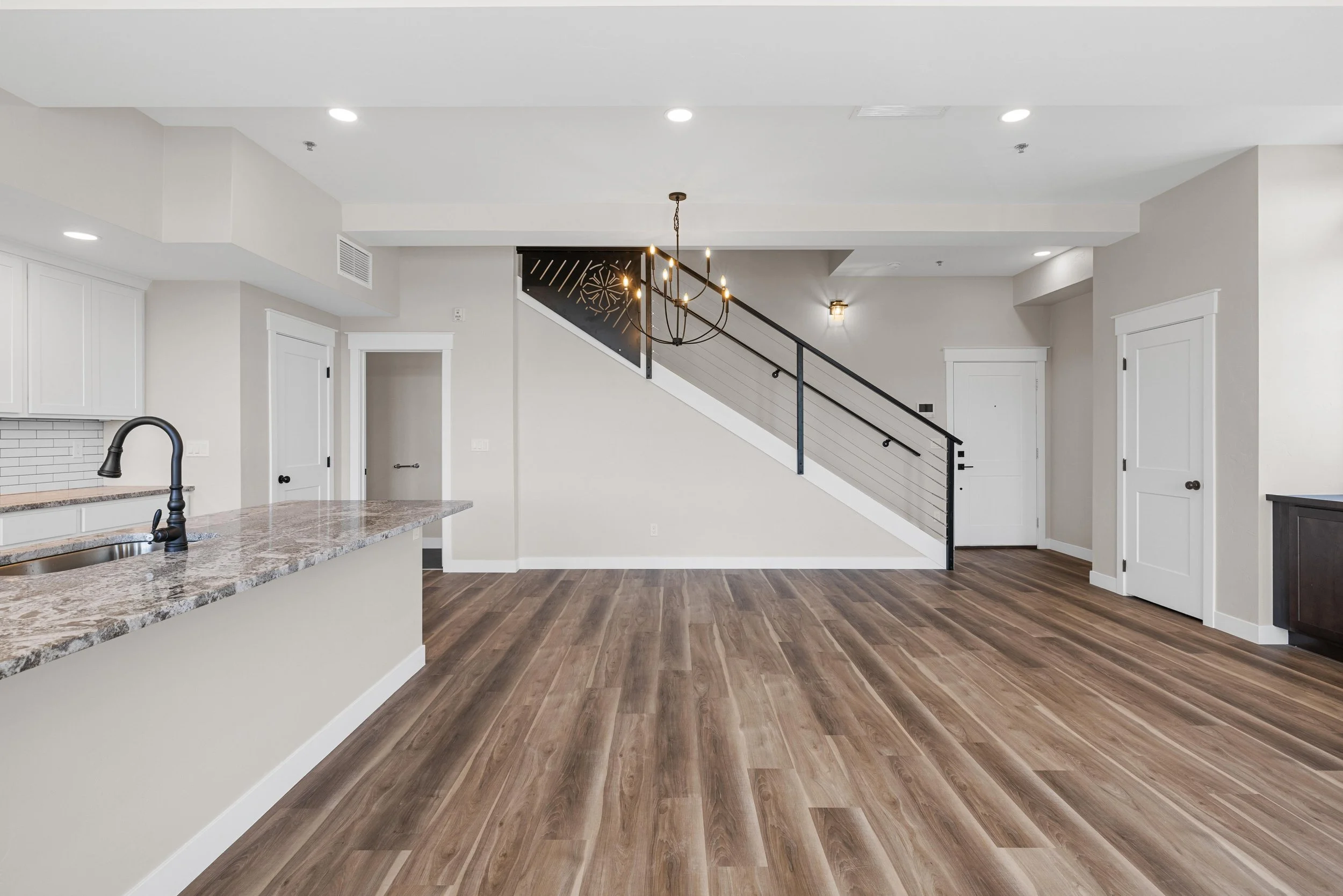 Empty living space with wood flooring, a kitchen island with a granite countertop, a staircase with black railing, white walls, and ceiling lights.