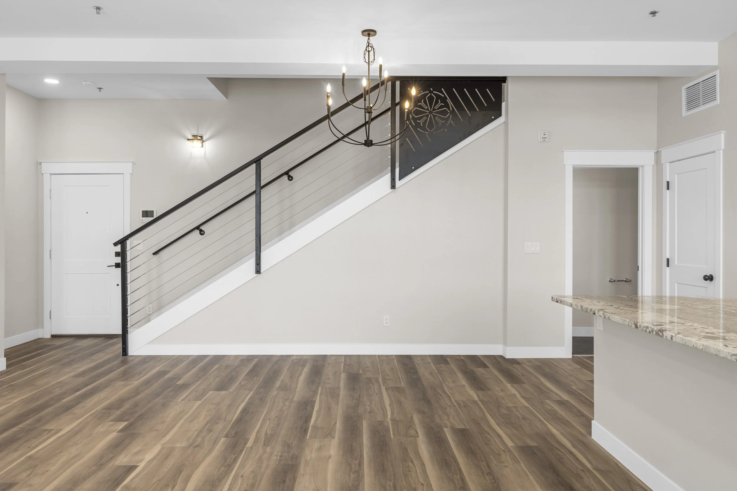 Empty living area with light wood flooring, white walls, a staircase with black railing, a chandelier, and partial view of a kitchen counter.