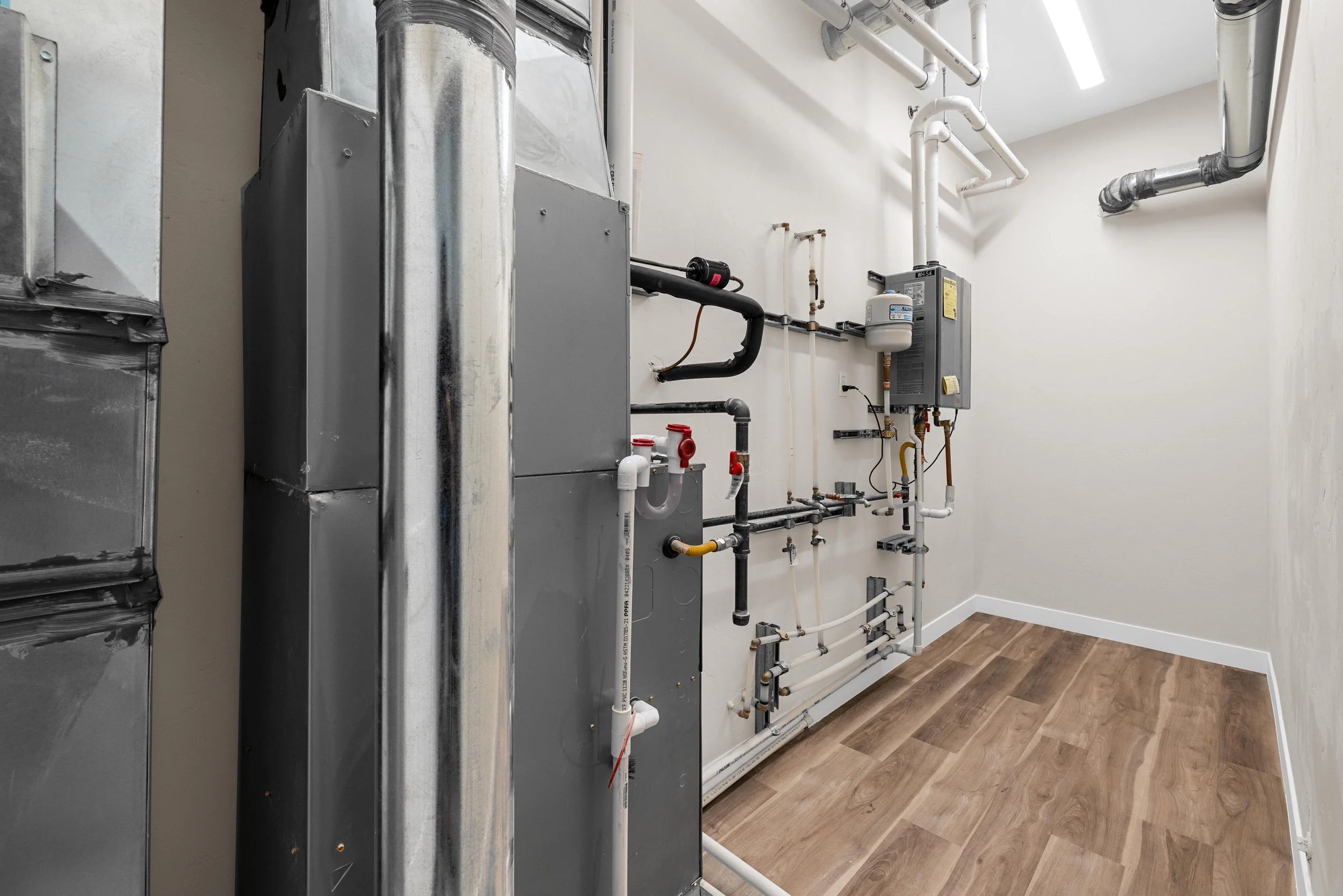 A utility room with various pipes, a water heater, and HVAC equipment mounted on a white wall, with a wood laminate floor.