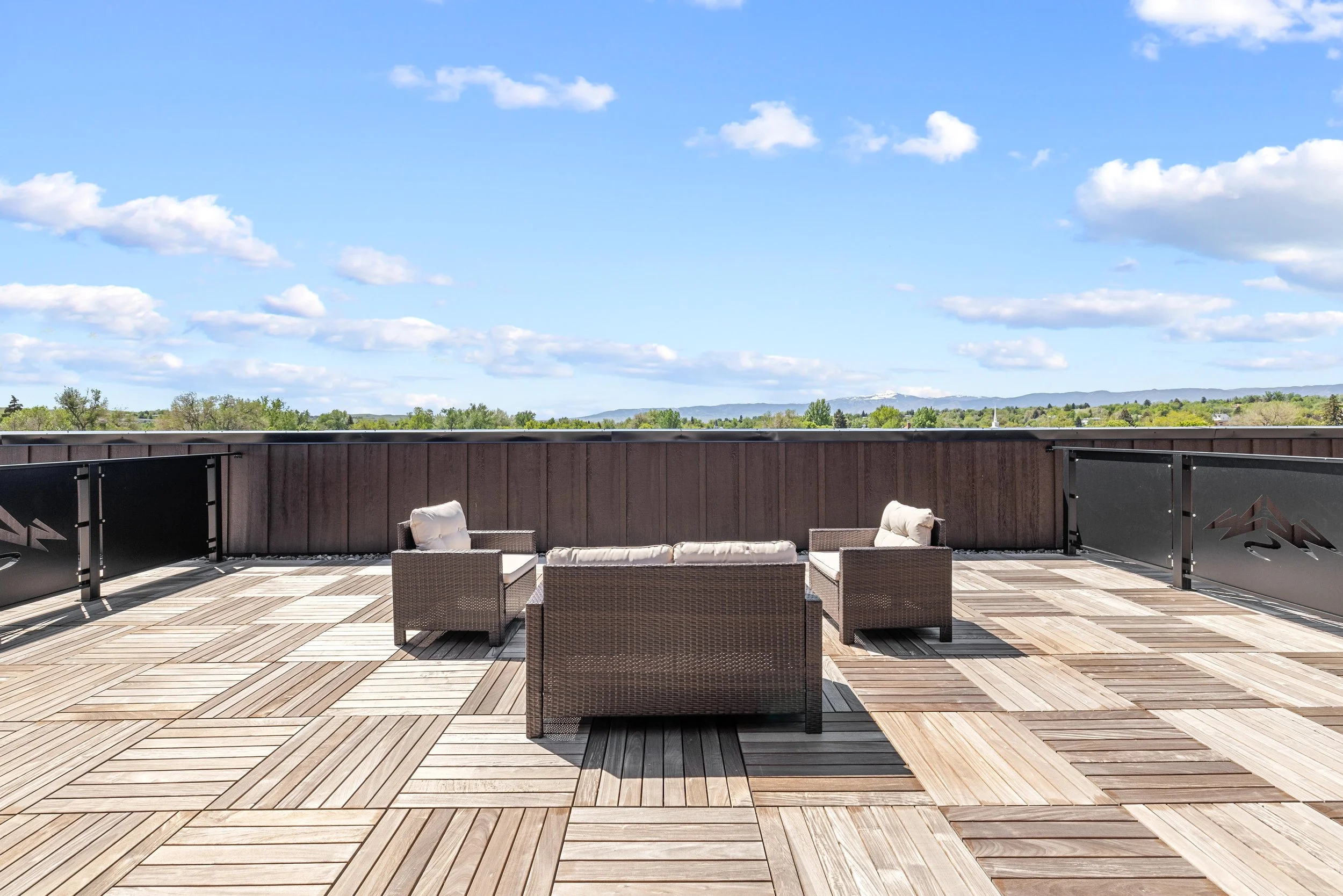 Rooftop patio with outdoor furniture, including a cushioned sofa and chairs, overlooking a landscape with trees and mountains under a blue sky with a few clouds.